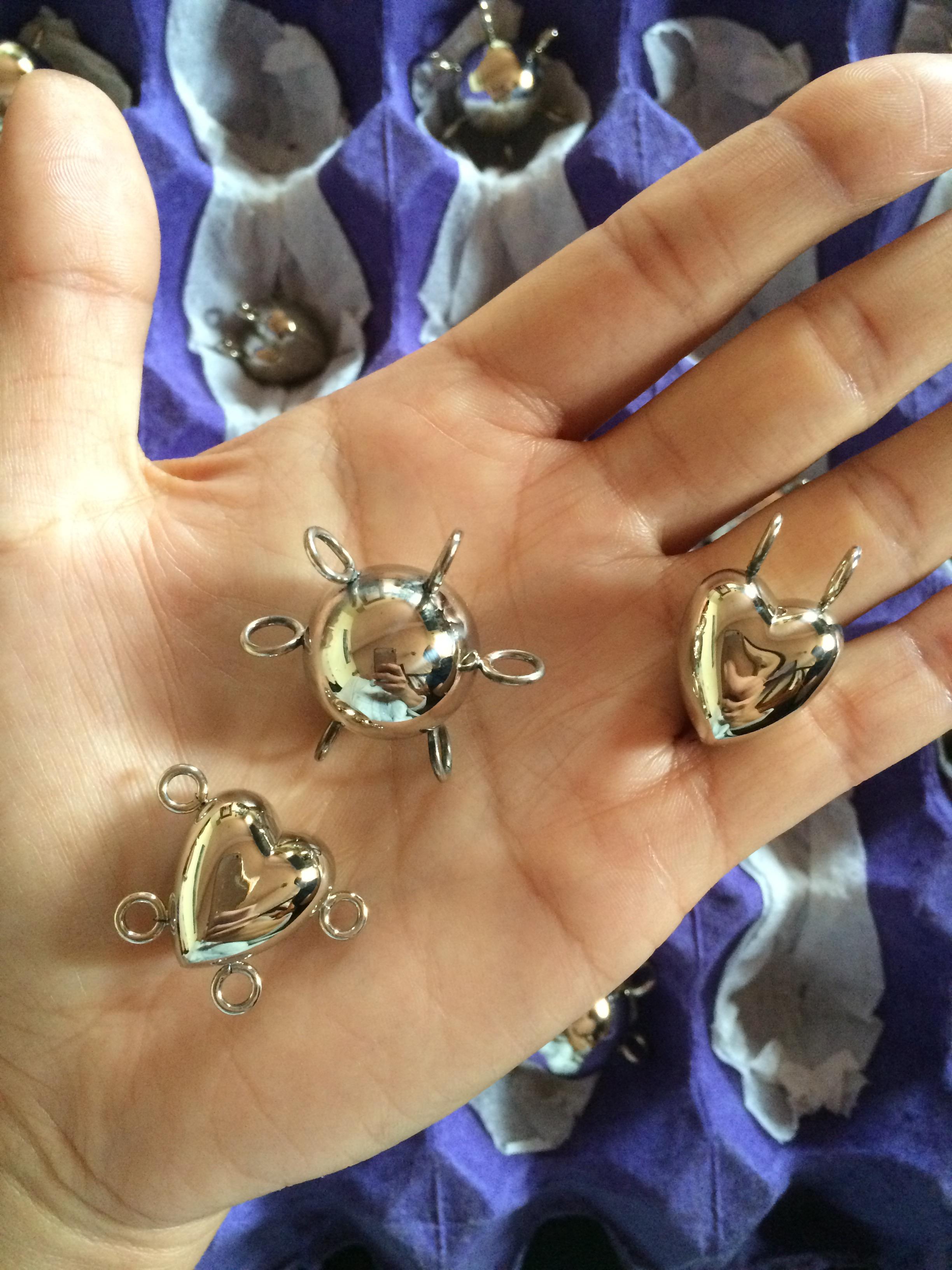 A close-up of a hand, palm up, holding three reflective silver charms. Two are heart-shaped and one is spherical, and each has several hooks for affixing to a chain.