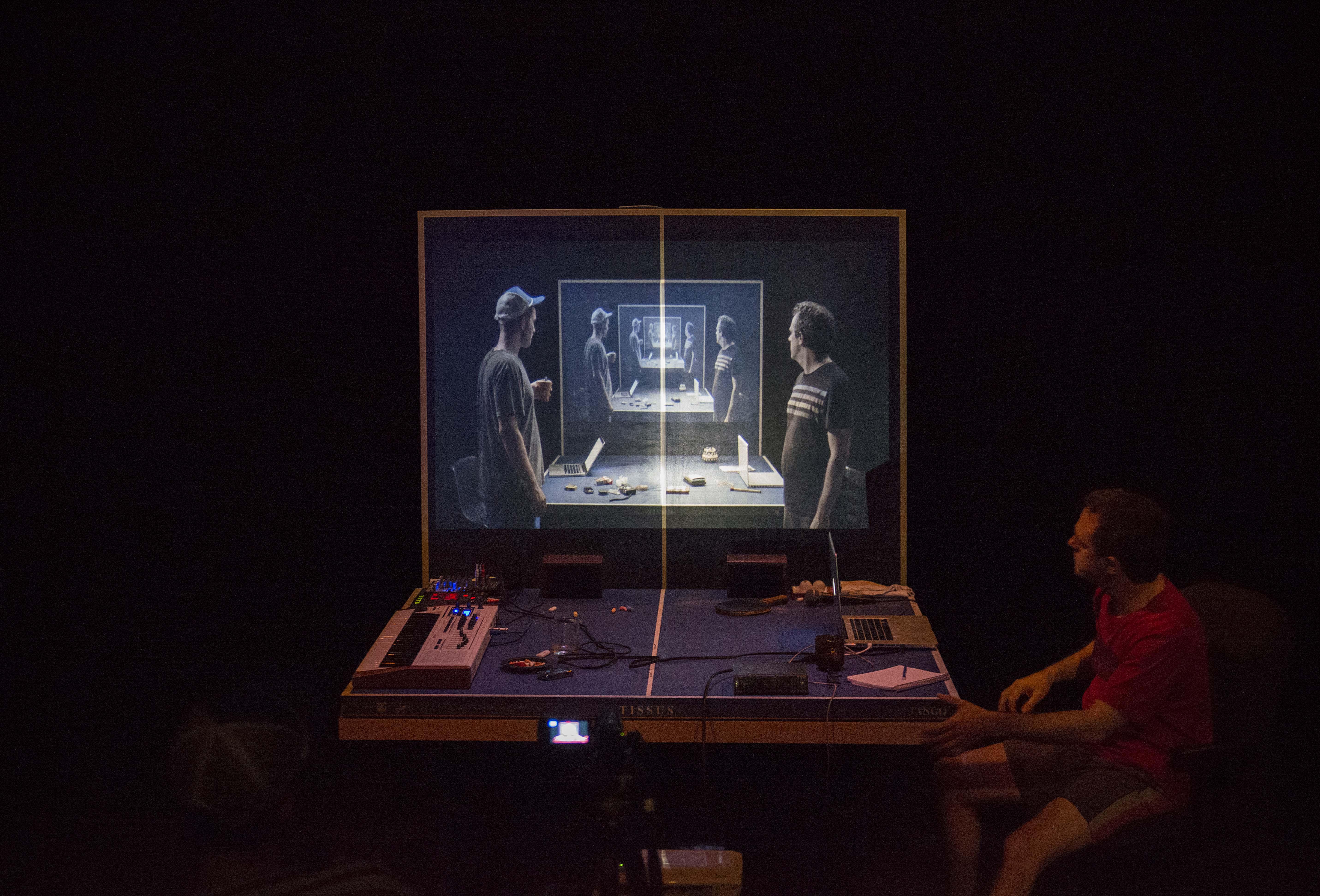 A man sits at a cluttered ping-pong table, looking at an image projected on the vertical rear half of the table. In the projected image, we see two men standing at the same table, their images repeating and receding into the distance.