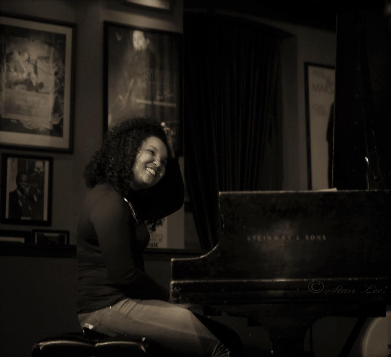 A woman sits at a piano in a dimly lit club. She tilts her head to the left and smiles.