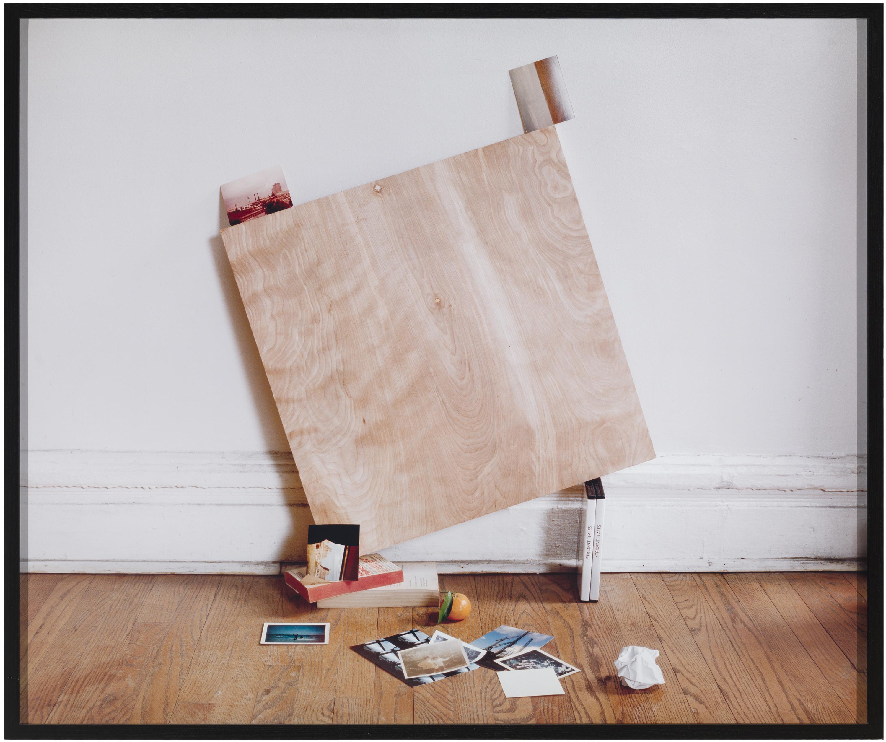 A square, wooden board precariously balanced on books leans against a white wall in a room with a hardwood floor scattered with photos.