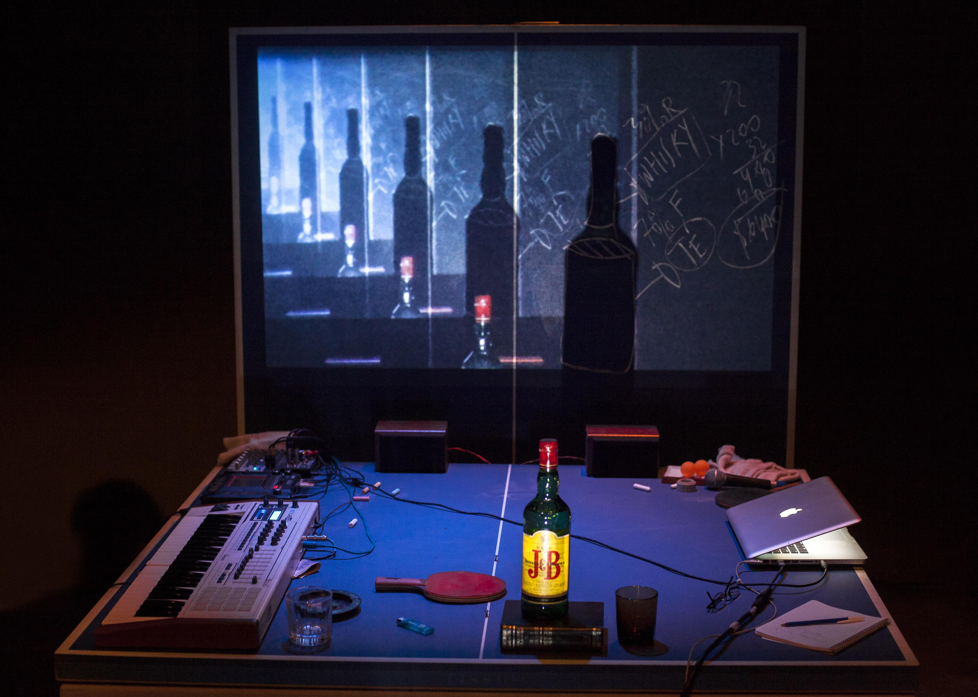 A ping-pong table is littered with a laptop, a musical keyboard, a bottle of whisky, glasses, cables, pens, and paper. The rear half of the table is vertical, showing a projected image of the bottle and its shadow that repeats and recedes into the distance.