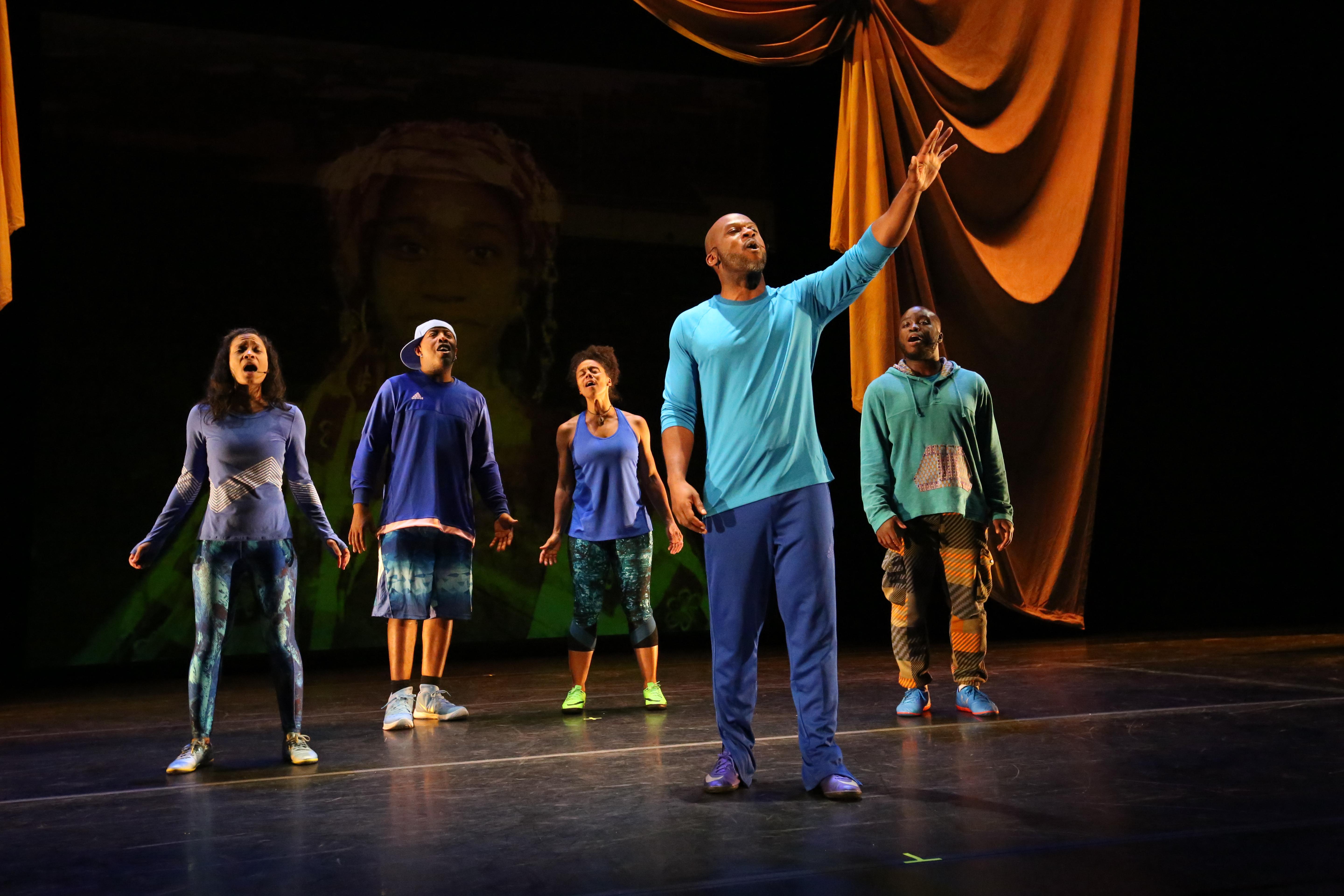 Five performers stand on a stage heads upraised and mouths open. They are dressed in athletic shoes and clothes in shades of blue. In the background, an elaborate umber drapery frames a faintly projected image of a young girl.