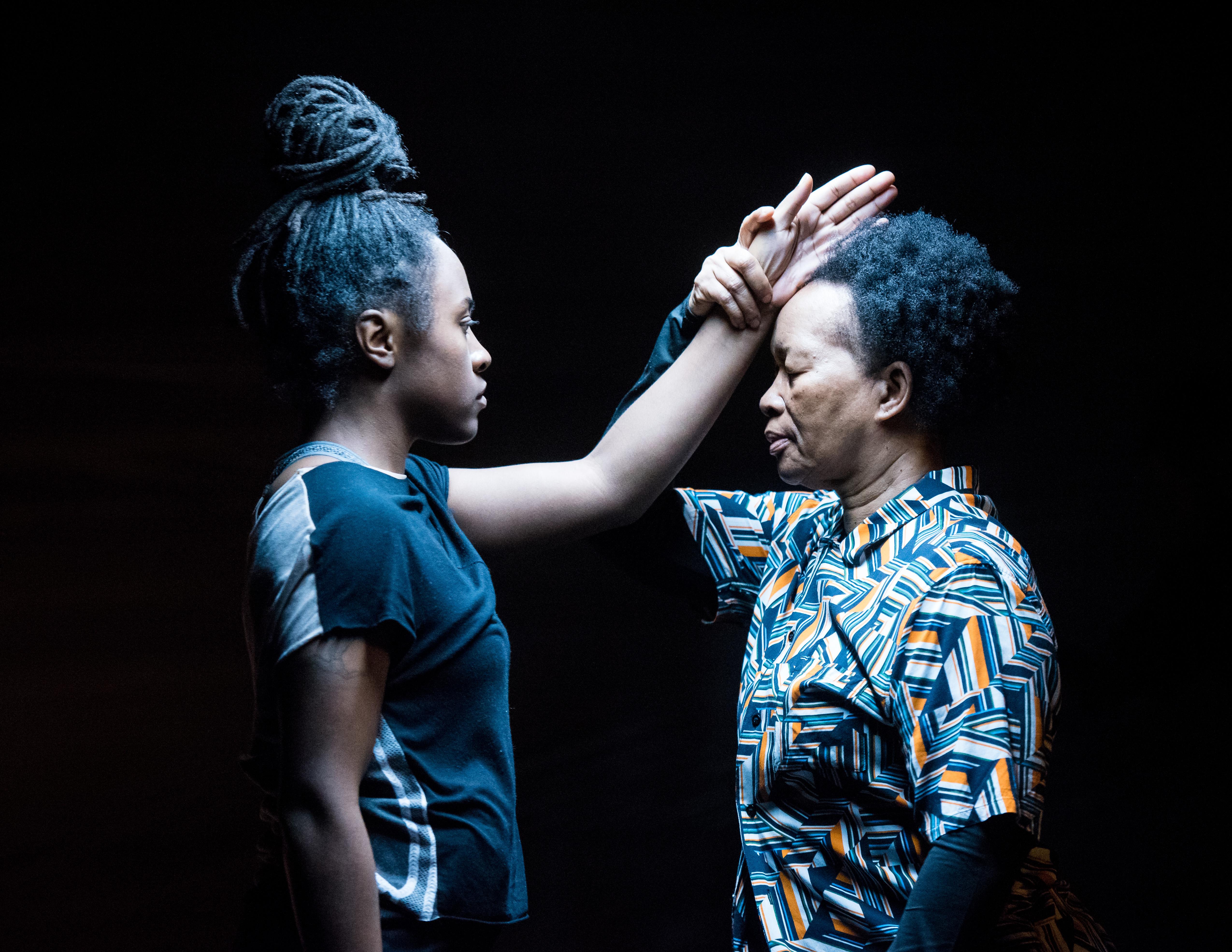 Two black women face one another eyes closed under a harsh fluorescent light. The older woman on the right holds the younger woman's outstretched arm by the wrist at her temple.