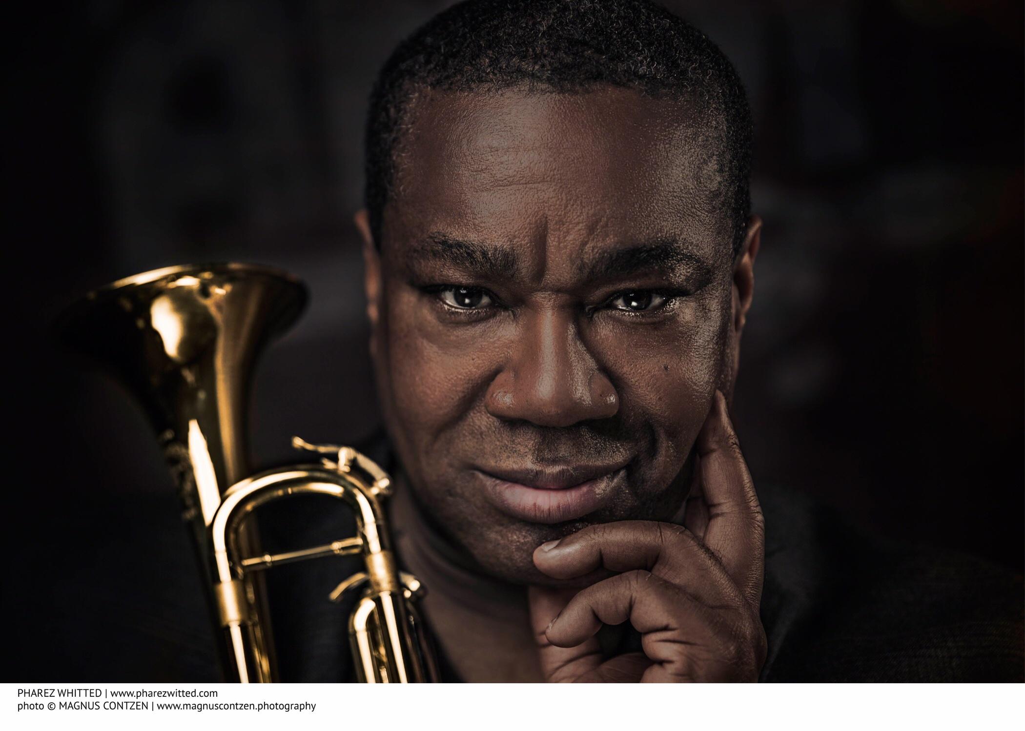 A black man smiles, posing with the bell end of a trumpet on his right. He rests his face on his left hand and looks directly at the camera, his brow furrowed.