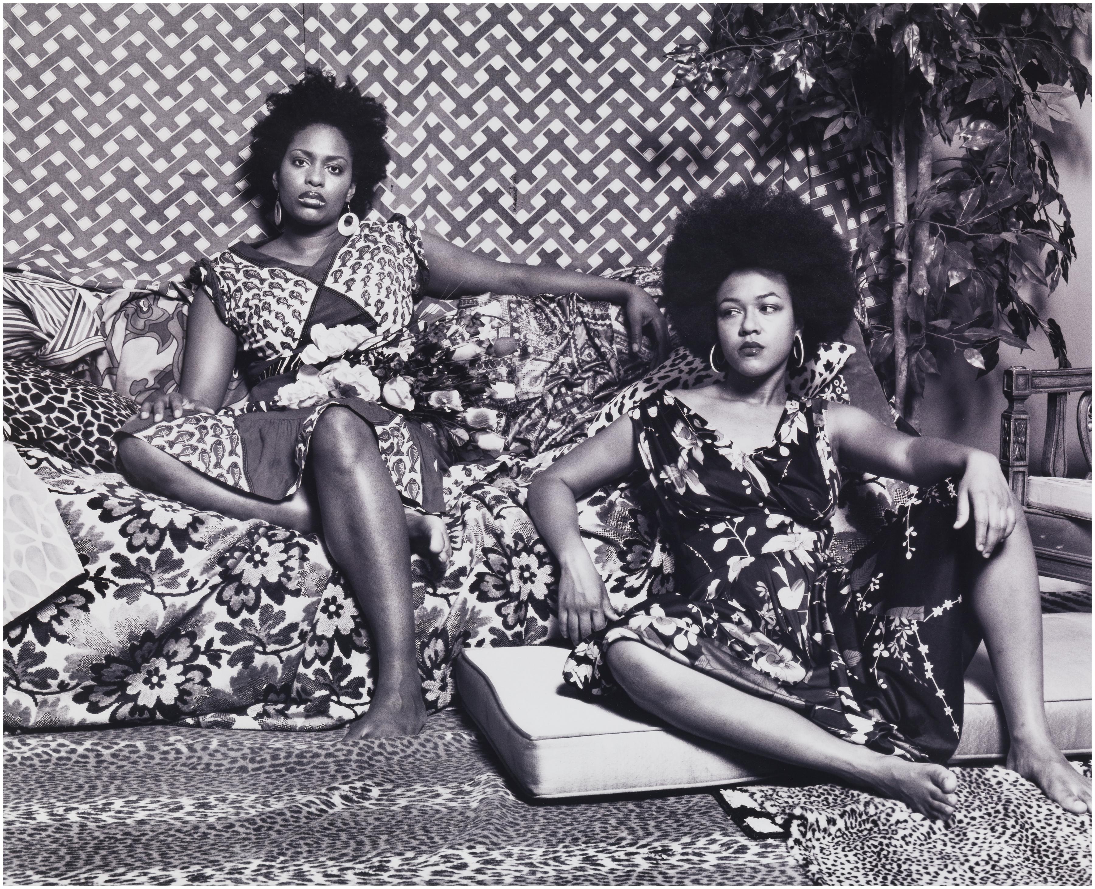 Two Black woman sit—one on a couch, one on a cushion on the floor—in a room filled with fabrics and rugs of many different patterns.