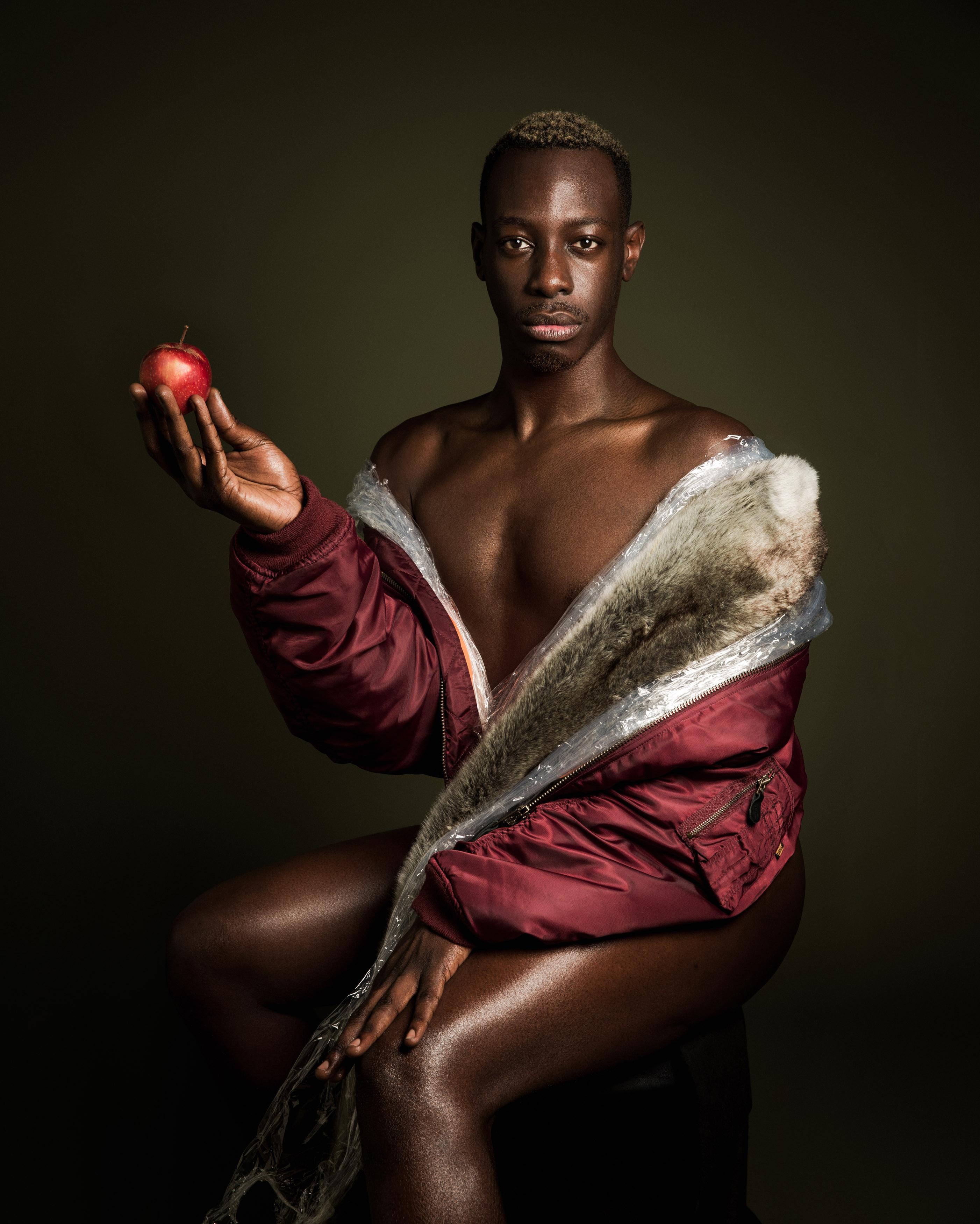 A dark-skinned person wears only a metallic, maroon, fur-lined jacket while seated, looking directly at the viewer and holding an apple in their right hand.