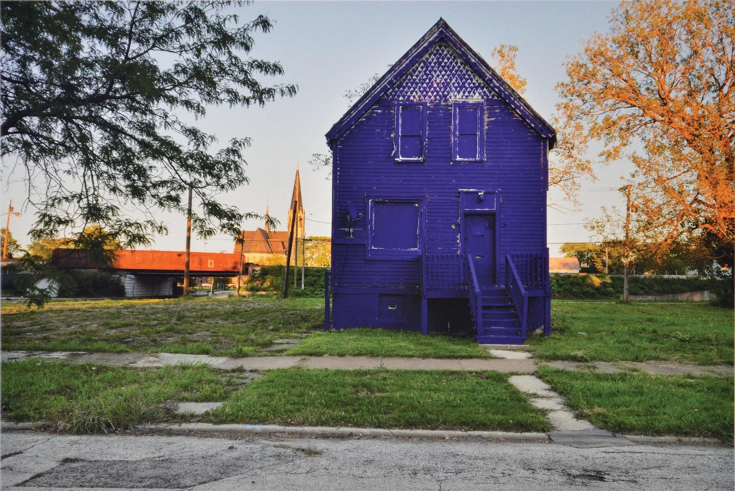 A two-story boarded up house is painted entirely blue. It is foregrounded by a patchy green lawn, old sidewalk, and damaged road.