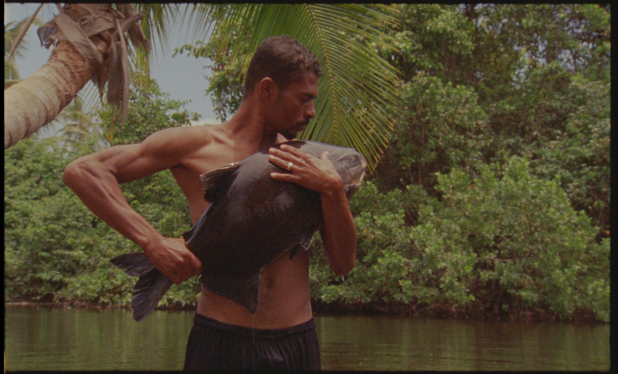 A shirtless person gently holds a fish against their body next to a river.