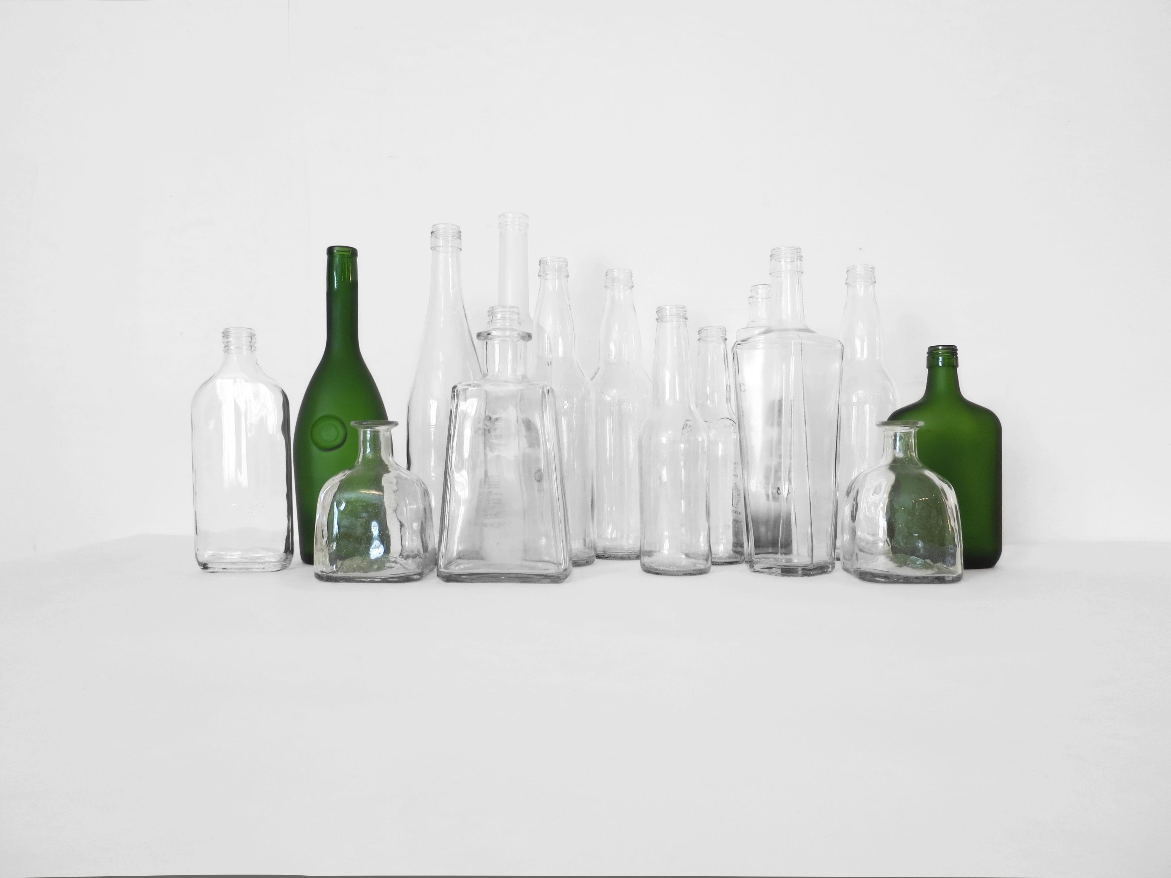 Thirteen colorless glass bottles and two green bottles, each a different shape and size and without a label, stand in a cluster in front of a white wall.