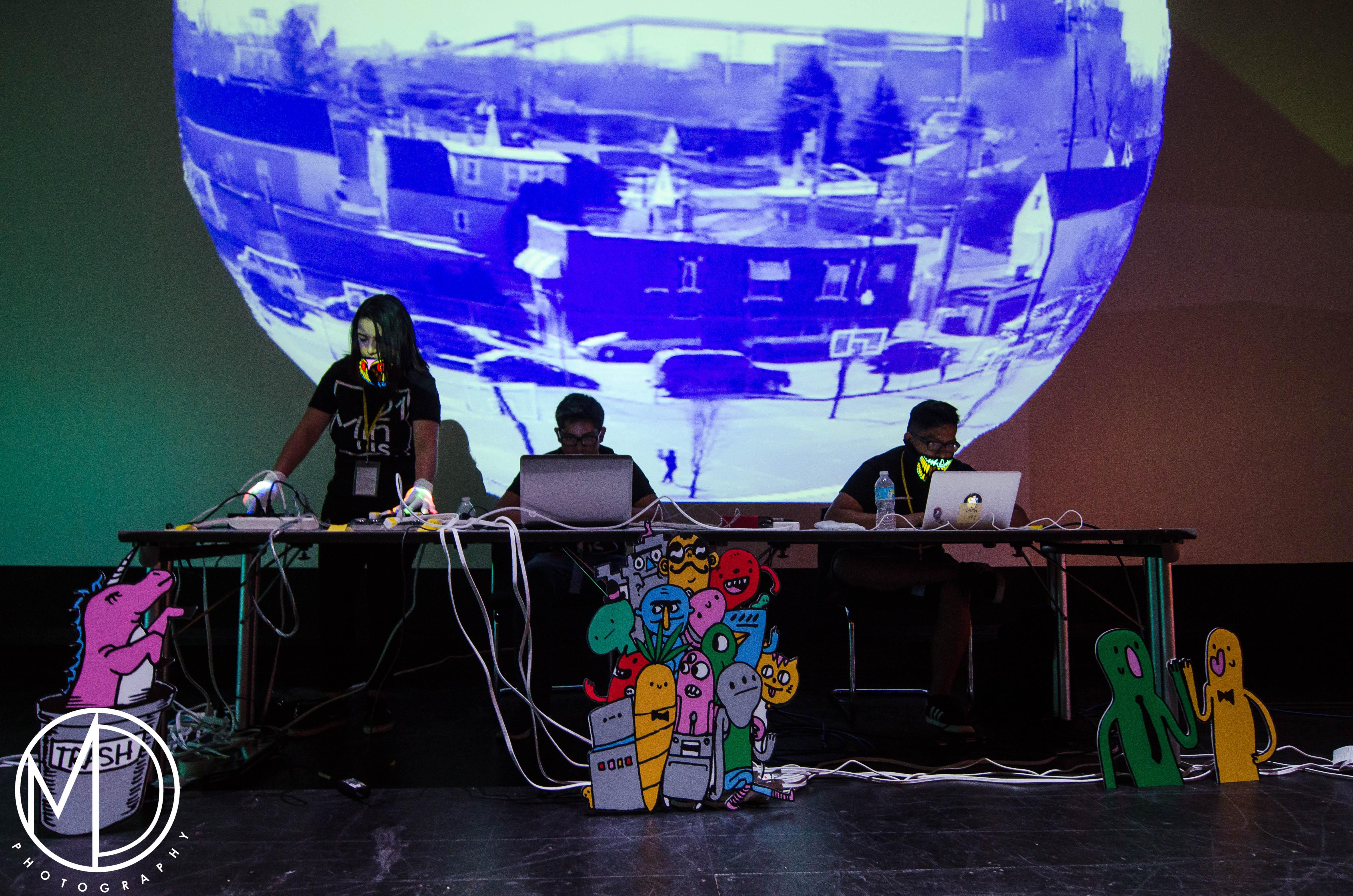 A projected sphere of a warped city landscape is foregrounded by three people at tables with laptops and electronics.