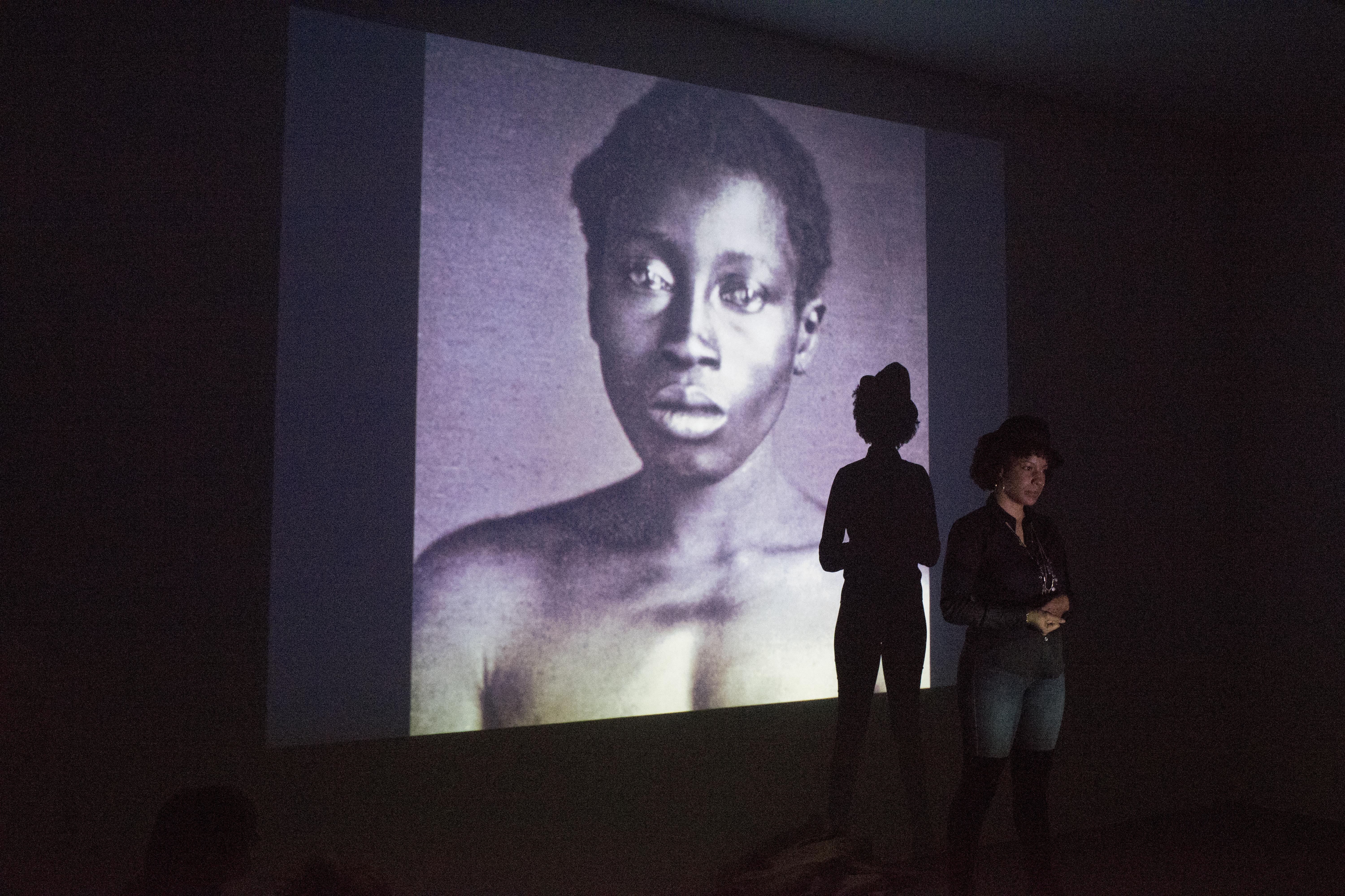 A black woman stands in front of a large projection of a topless, black woman.