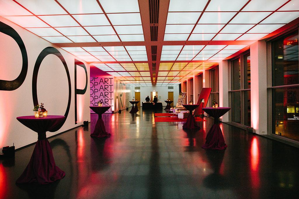 A long room is set up for a party with red uplighting on the walls and cocktail tables wrapped in purple fabric lining each side of the room.