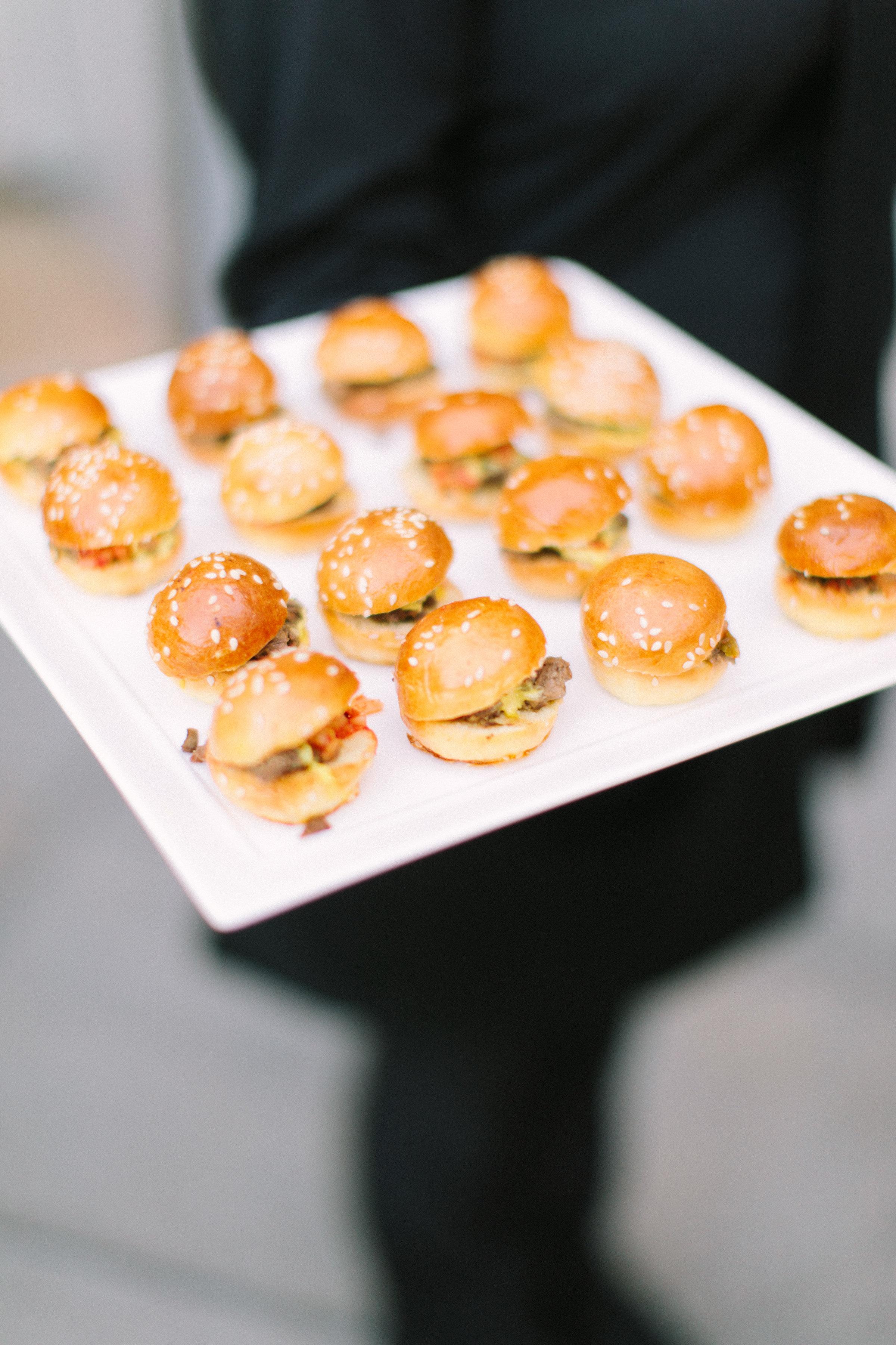 a server holds a plate of hor d'oeuvres in the form of small sandwiches on golden sesame buns
