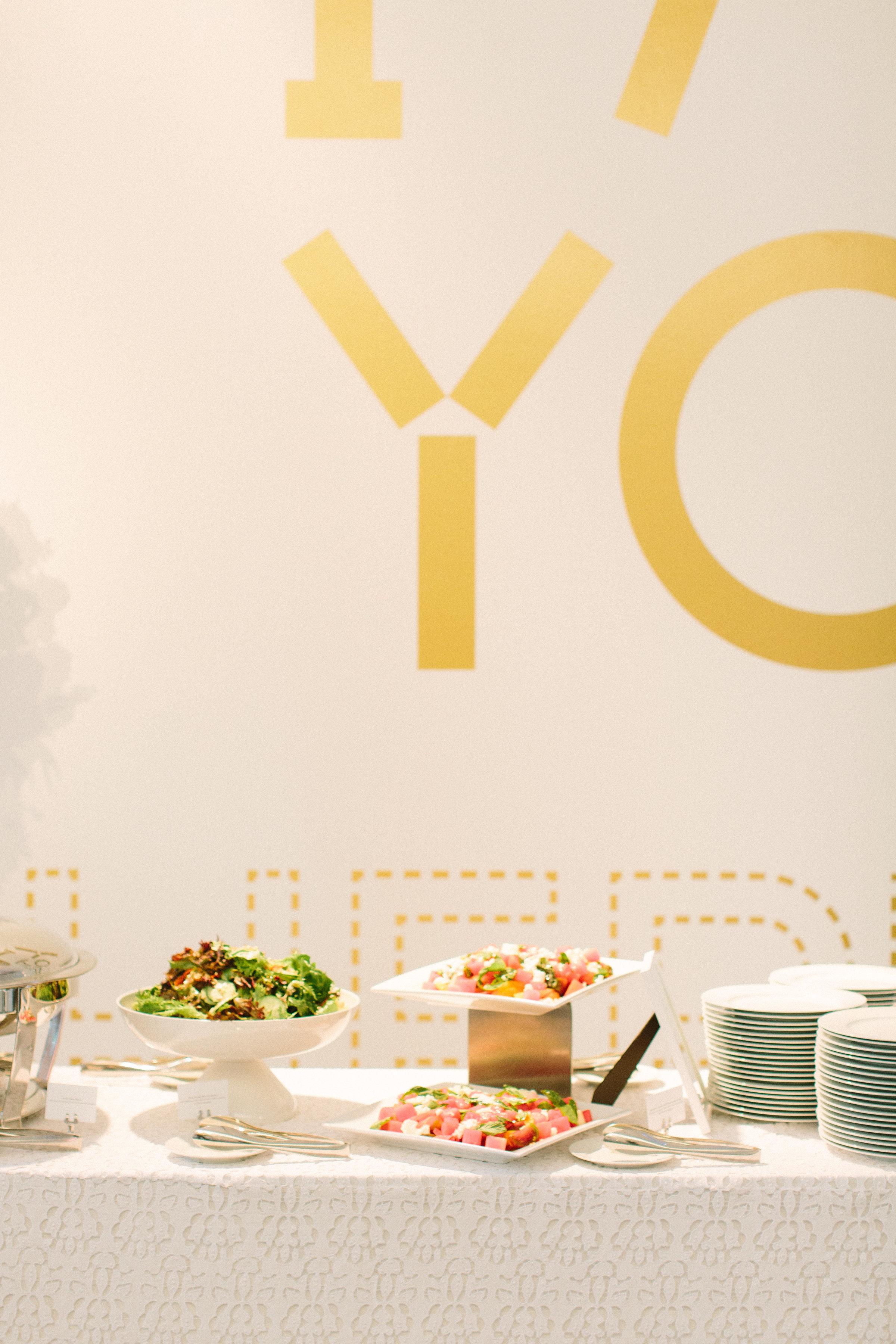 An elegant display of green and watermelon salads are displayed on white tableware besides stacked white plates on a contemporary-patterned white tablecloth.