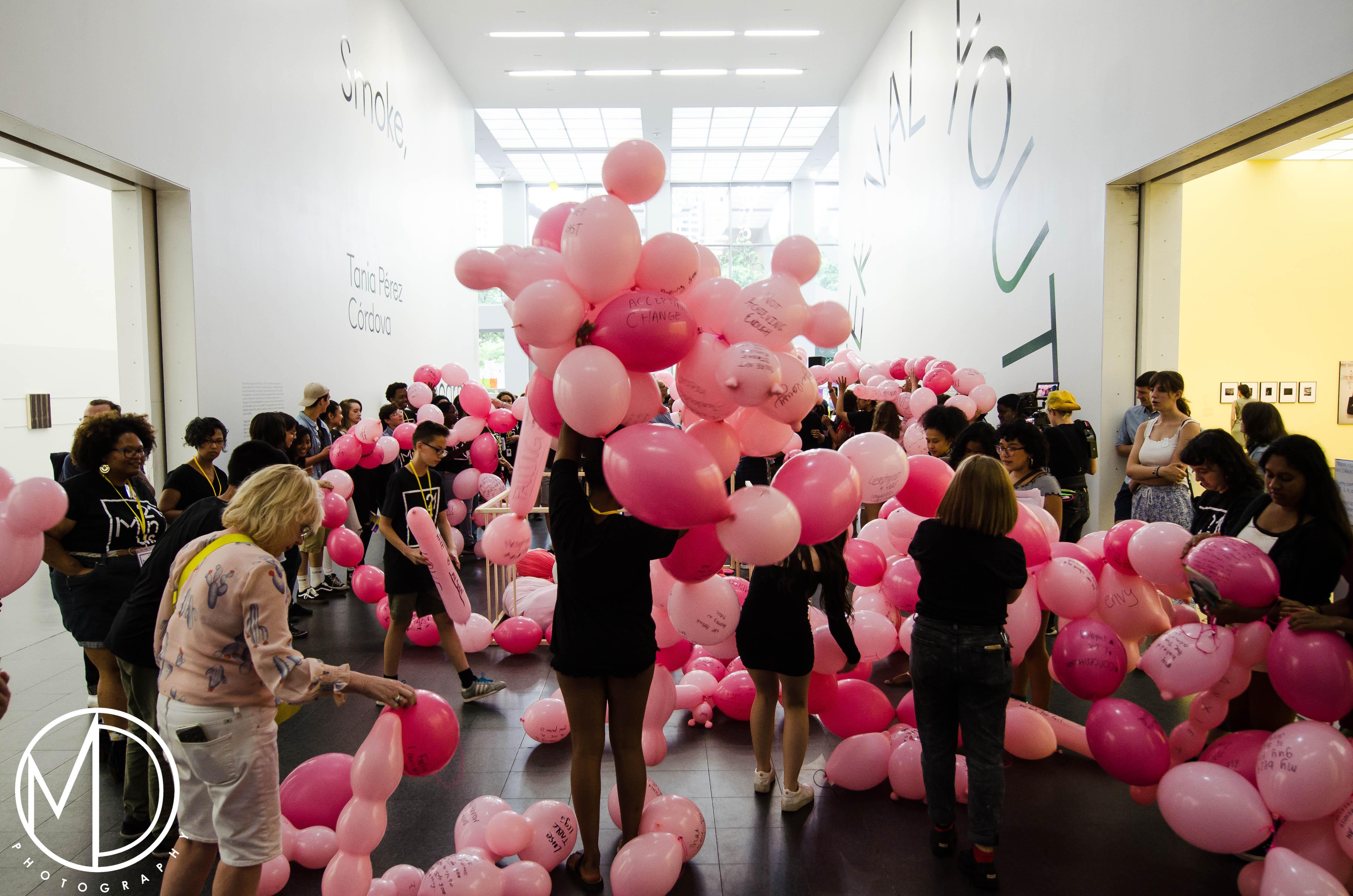 A young person's back is to us as they lift a cluster of pink balloons over their head in the MCA's atrium. Surrounding them are a sea of balloons and people doing similar playful motions.