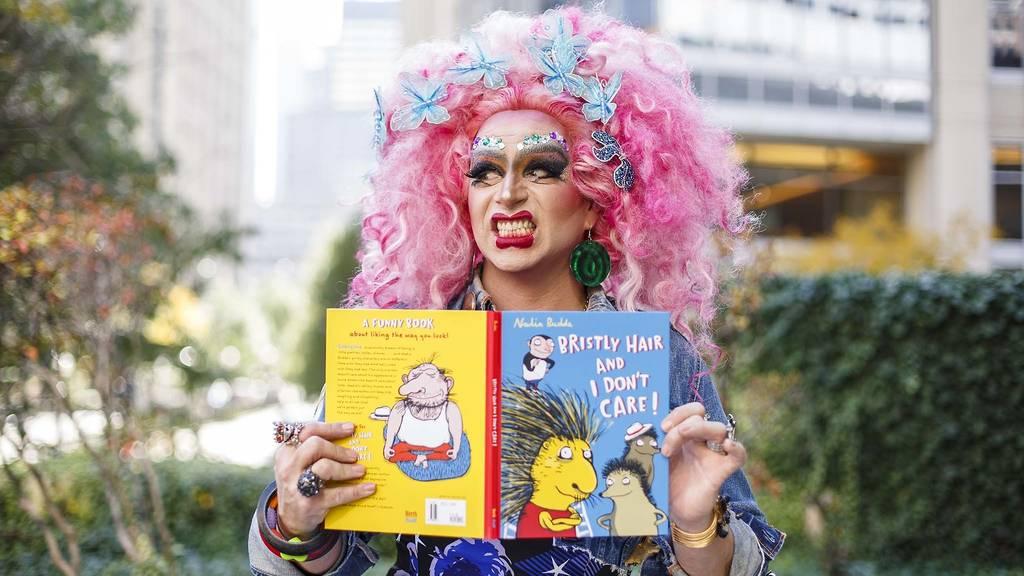 A drag queen in a pink wig looks up from the children's book she is holding.
