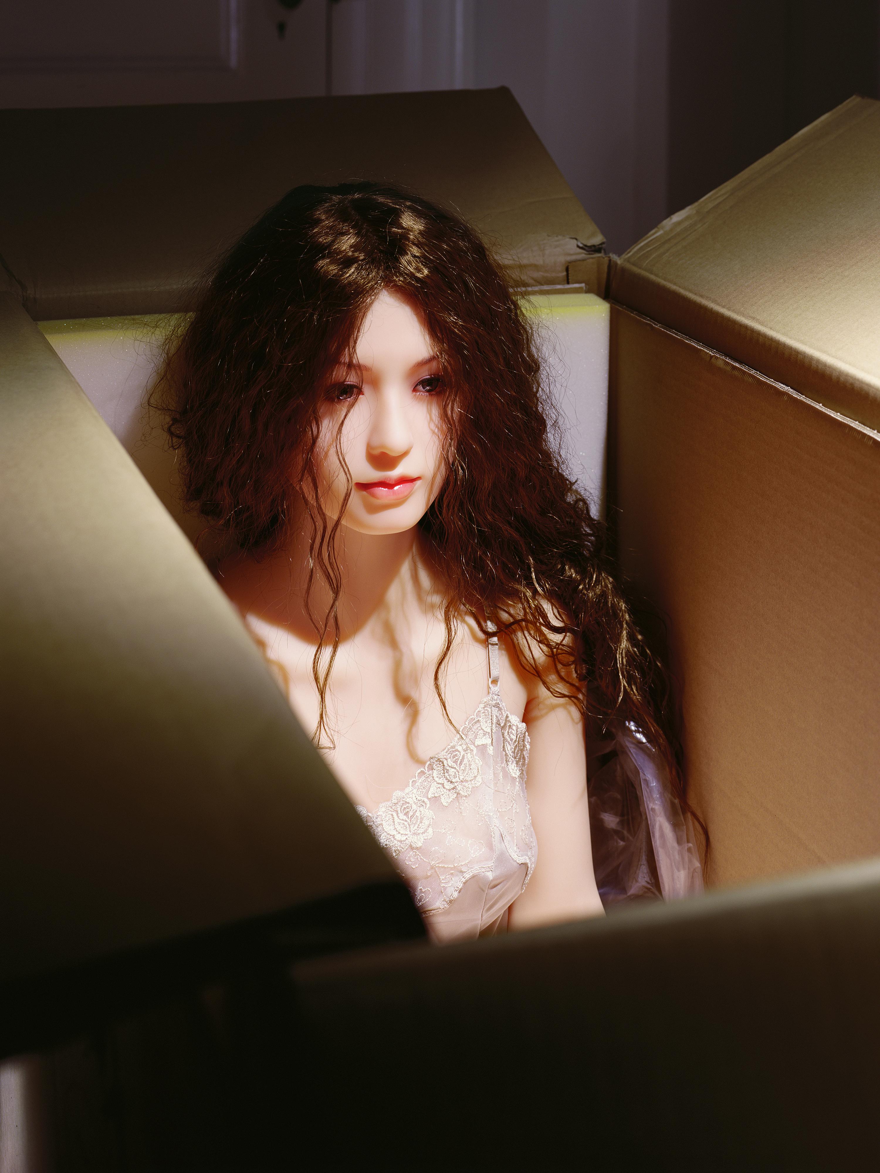 A feminine doll with long hair sits upright in a cardboard box. She wears a white negligee with lace details.