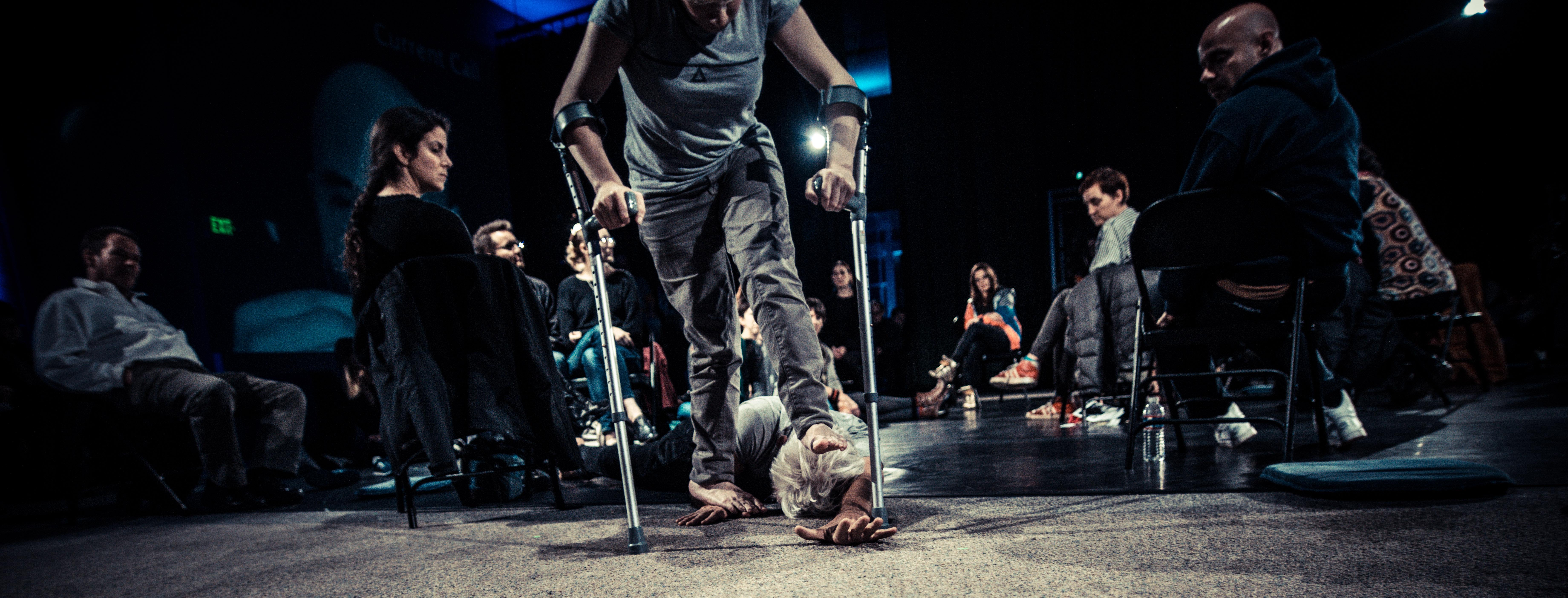 People seated on a darkened stage watch as a woman using crutches steps on and walks over a man, who lies on the ground.