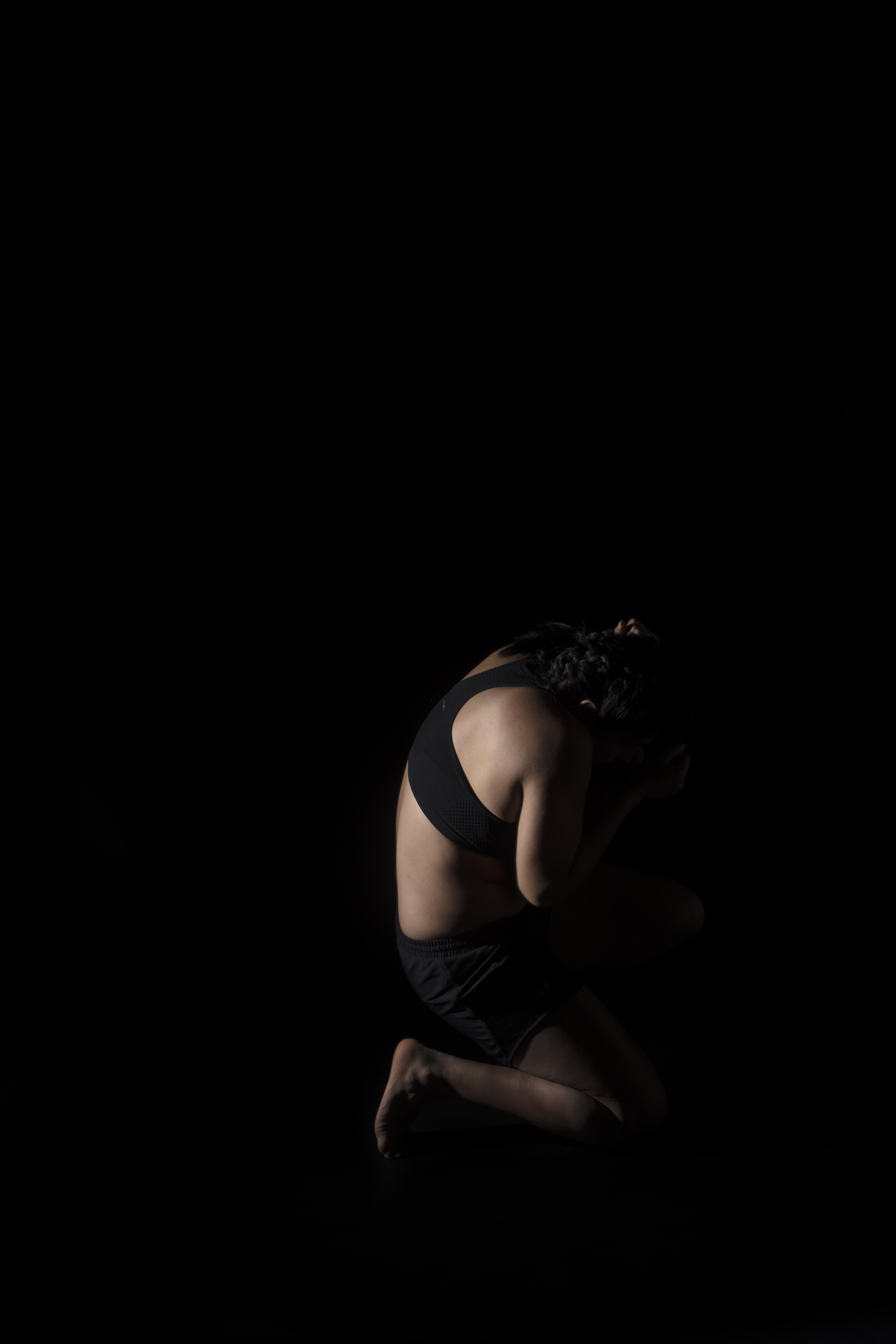 A person crouches in a dark space on one knee, sheilding their face from the light.