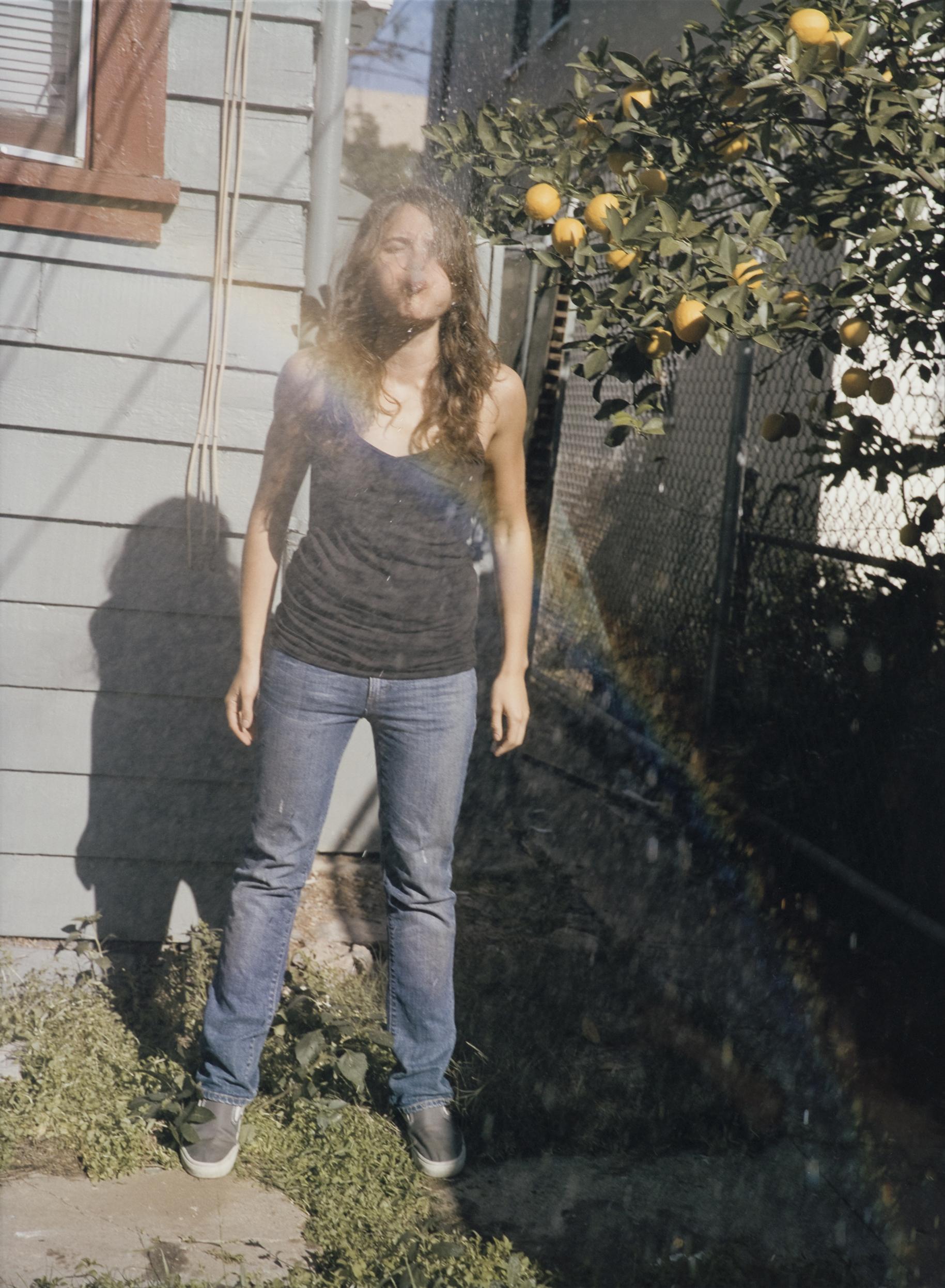 A young woman spits water at you, creating a rainbow through the spray. She stands in a backyard next to a lemon tree.