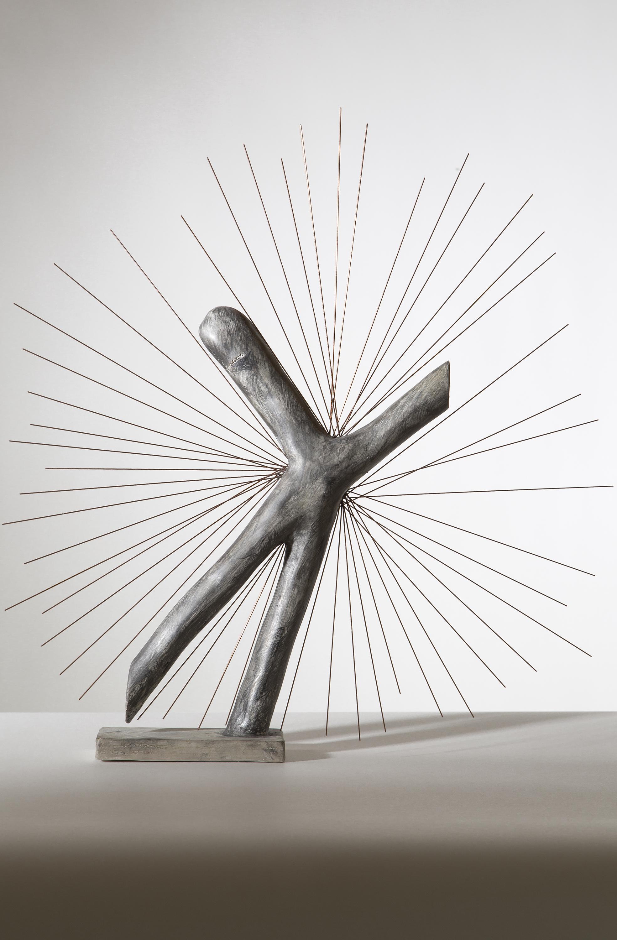 A sculpture shaped like a crooked letter X stands on a short rectangular base. From the center many spokes jut out creating a starburst effect.