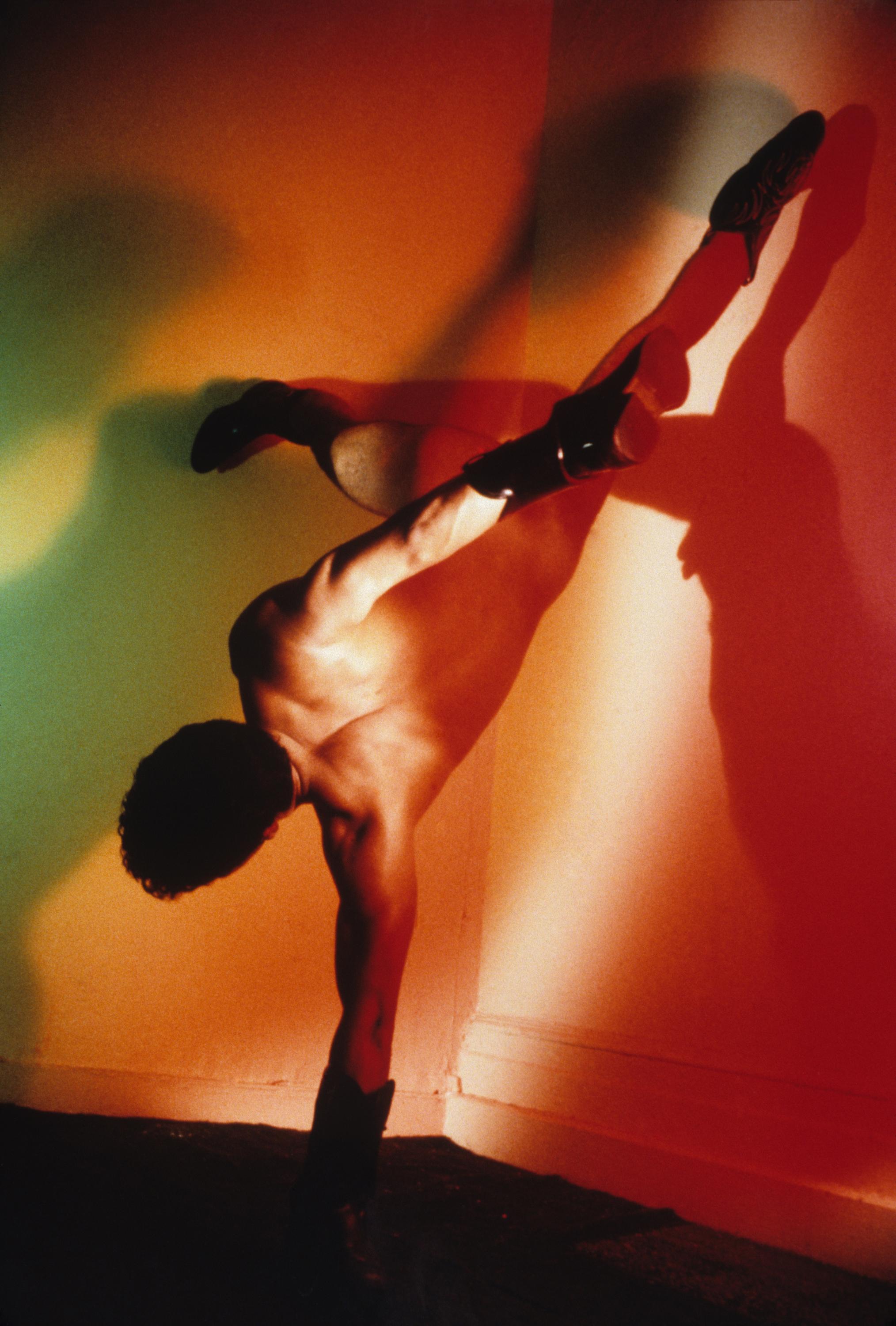 A nude adult with masculine shoulders wears cowboy boots on their hands and feet. They stand on one of their hands with their feet splayed across the corner of a room lit in red.