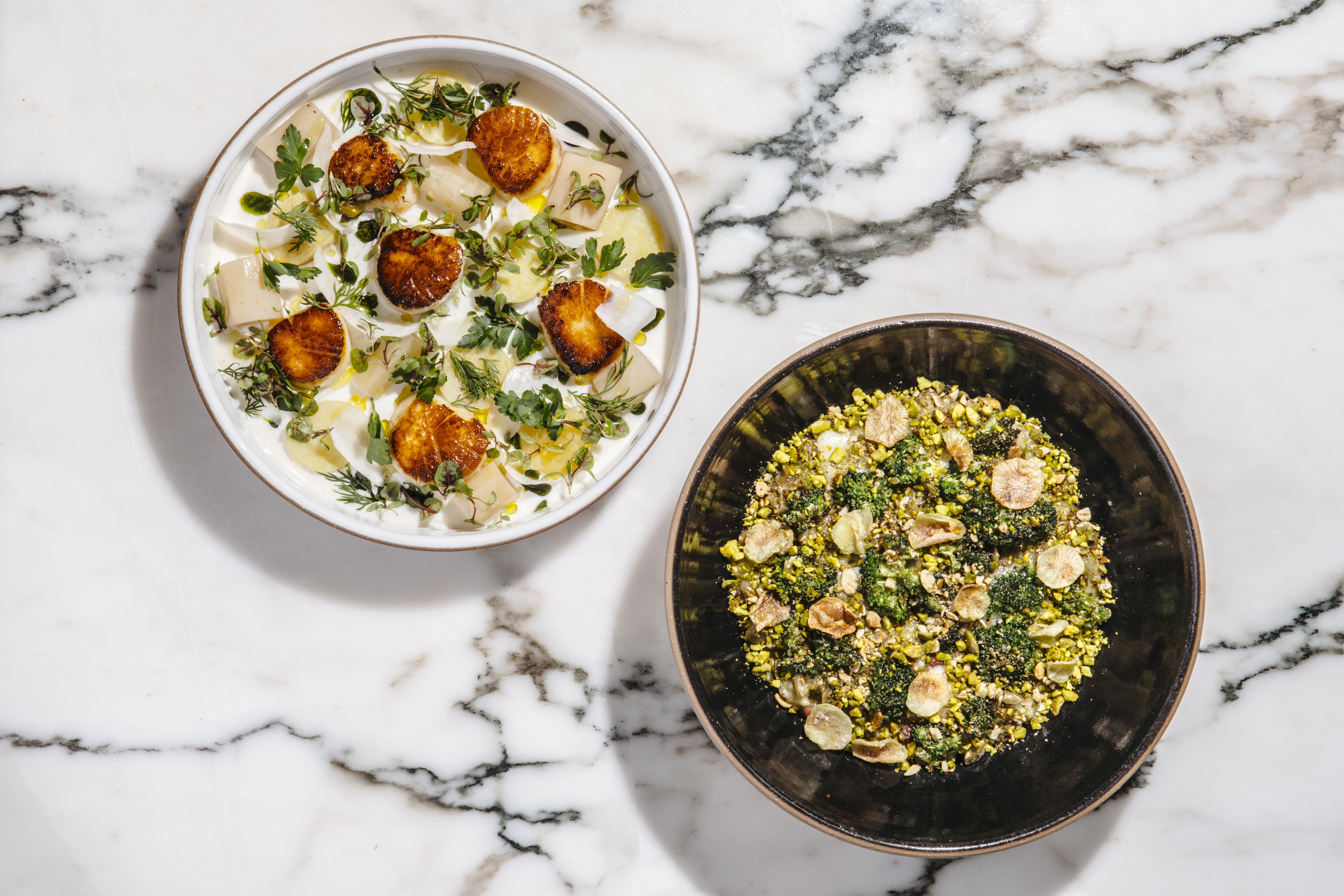 Two dishes are plated and served atop a white marble surface. The dish on the left is light, with greens and six scallops. The dish on the right appears more dense.