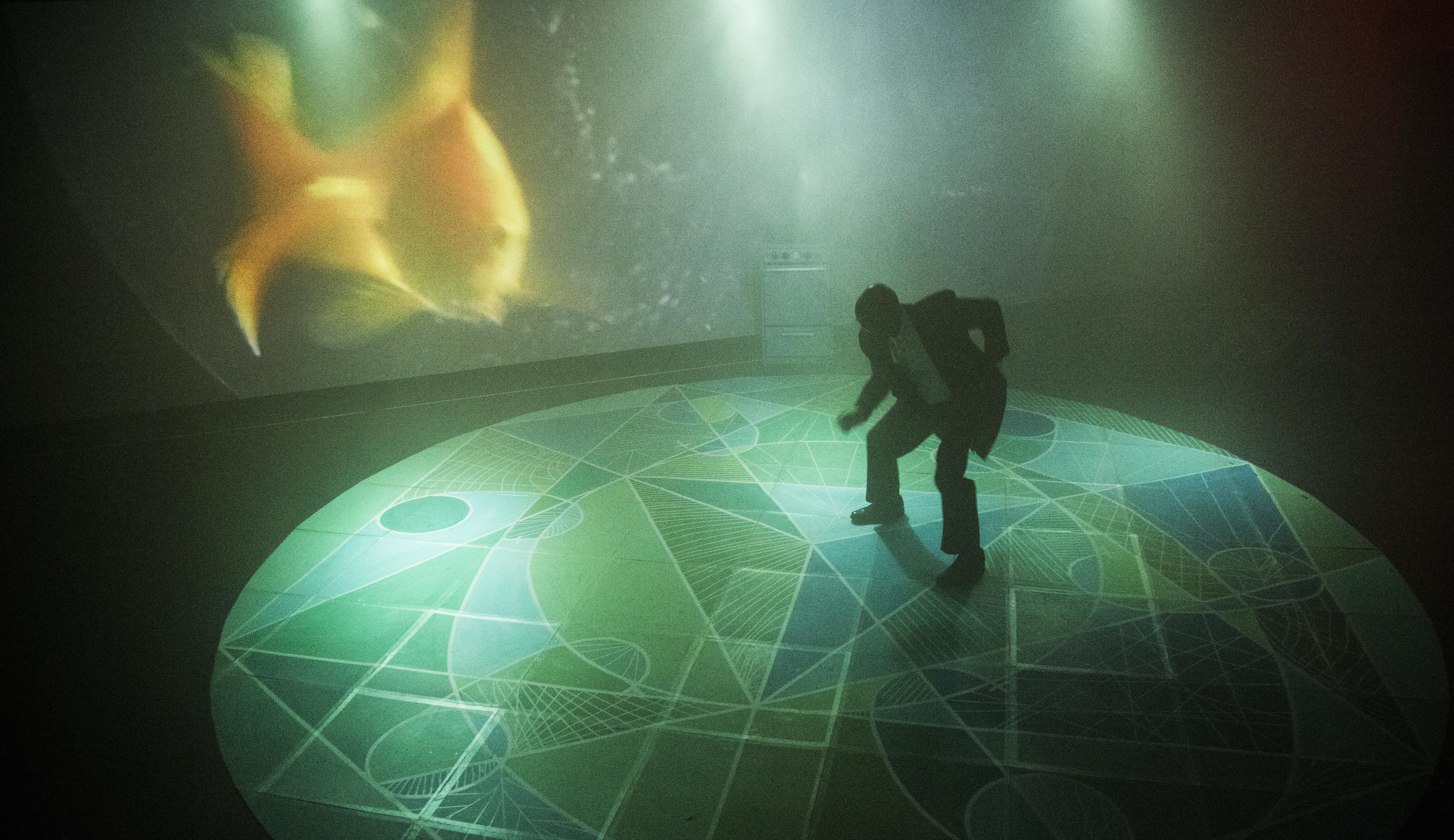 A man in a businesslike suit crouches as if dancing on an illuminated stage. The floor beneath him features an intricate pattern of various greens, while the screen behind him shows a blurry projection of a goldfish.