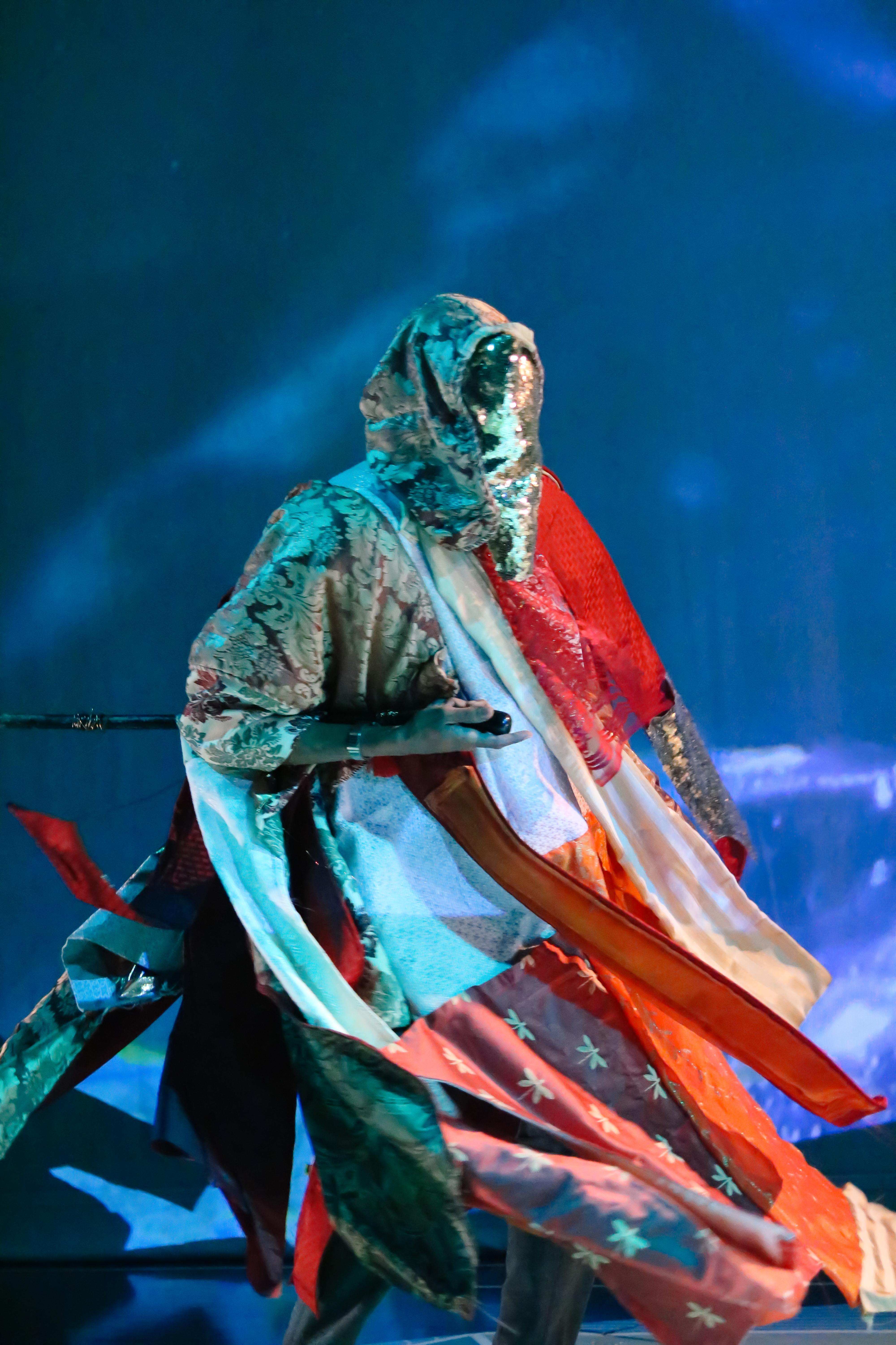 A masked figure is draped in a flowing, shimmering robe assembled from multiple gold, red, and white textiles.