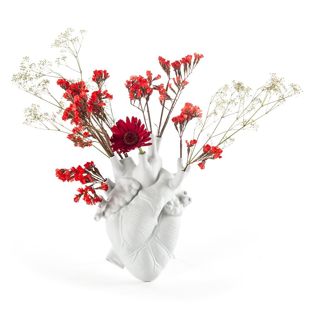 A porcelain vase in the shape of a human heart with red flowers arranged in the valve openings