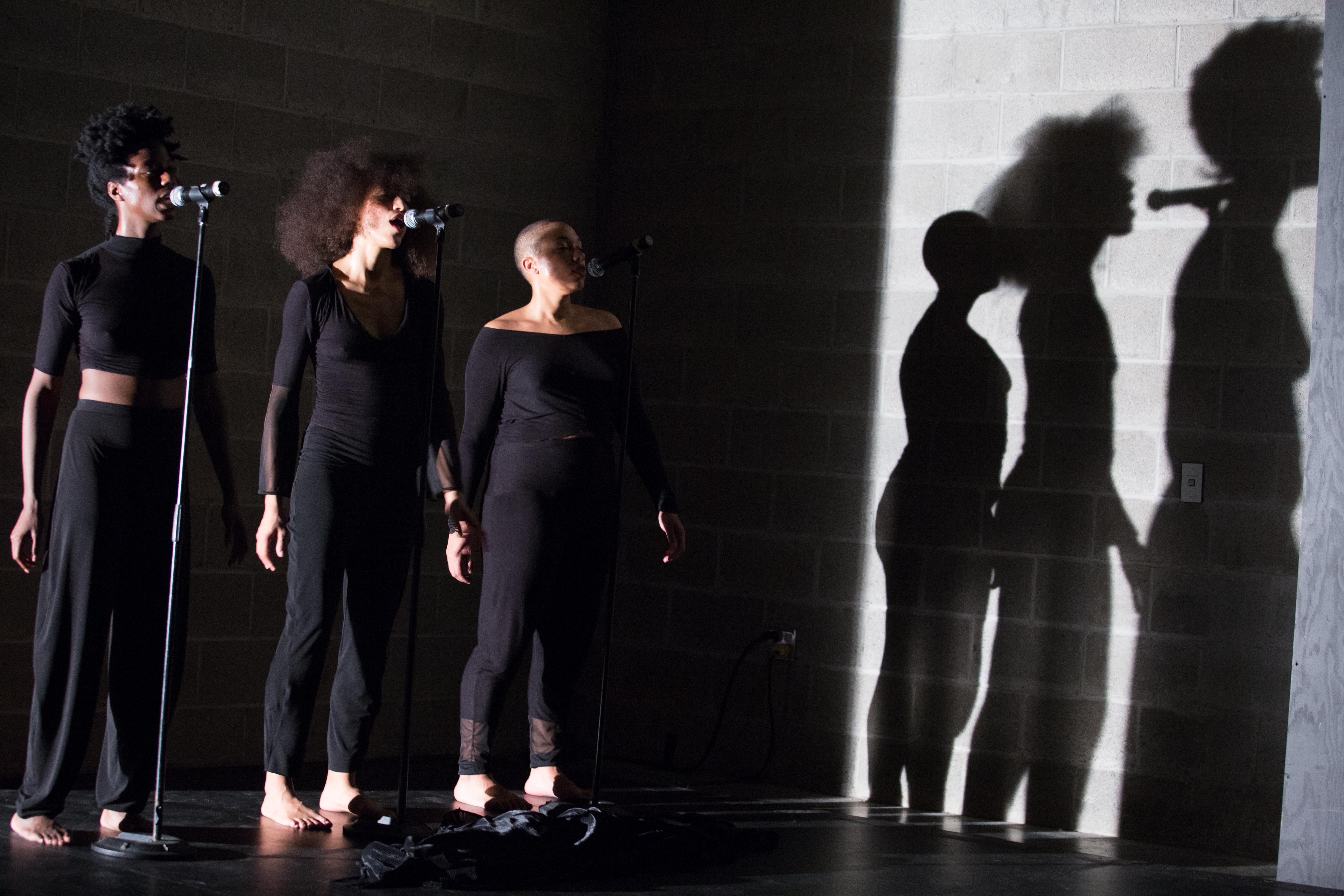 three performers stand spotlit at microphones with shadows cast in background