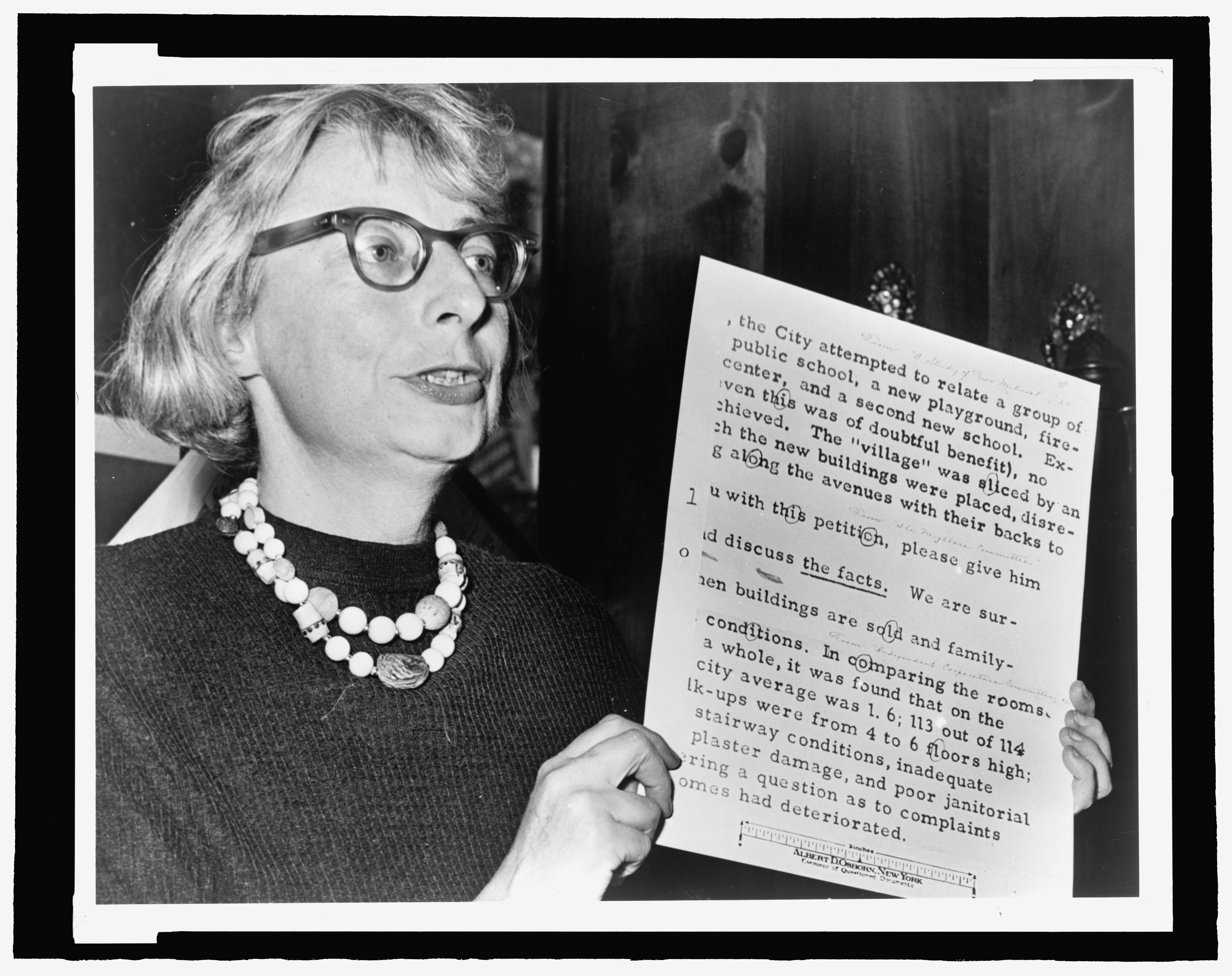 A black-and-white photograph shows a middle-aged, light-skinned woman holding a printed document aloft. It appears as though the woman is speaking.