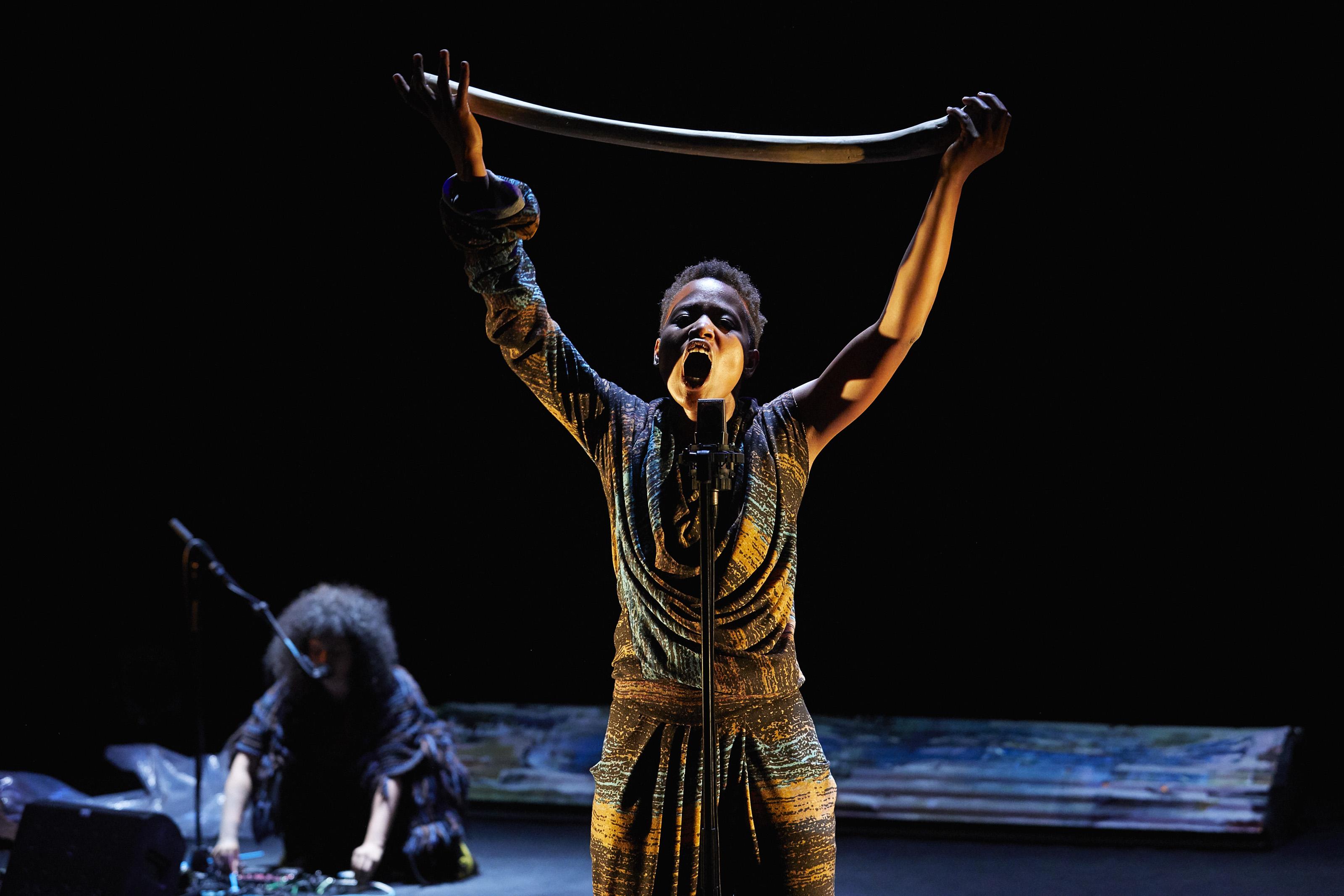 A young black woman stands at a microphone, her mouth open in a cry or in song. She holds a curved length of wood or bone above her head with both hands.