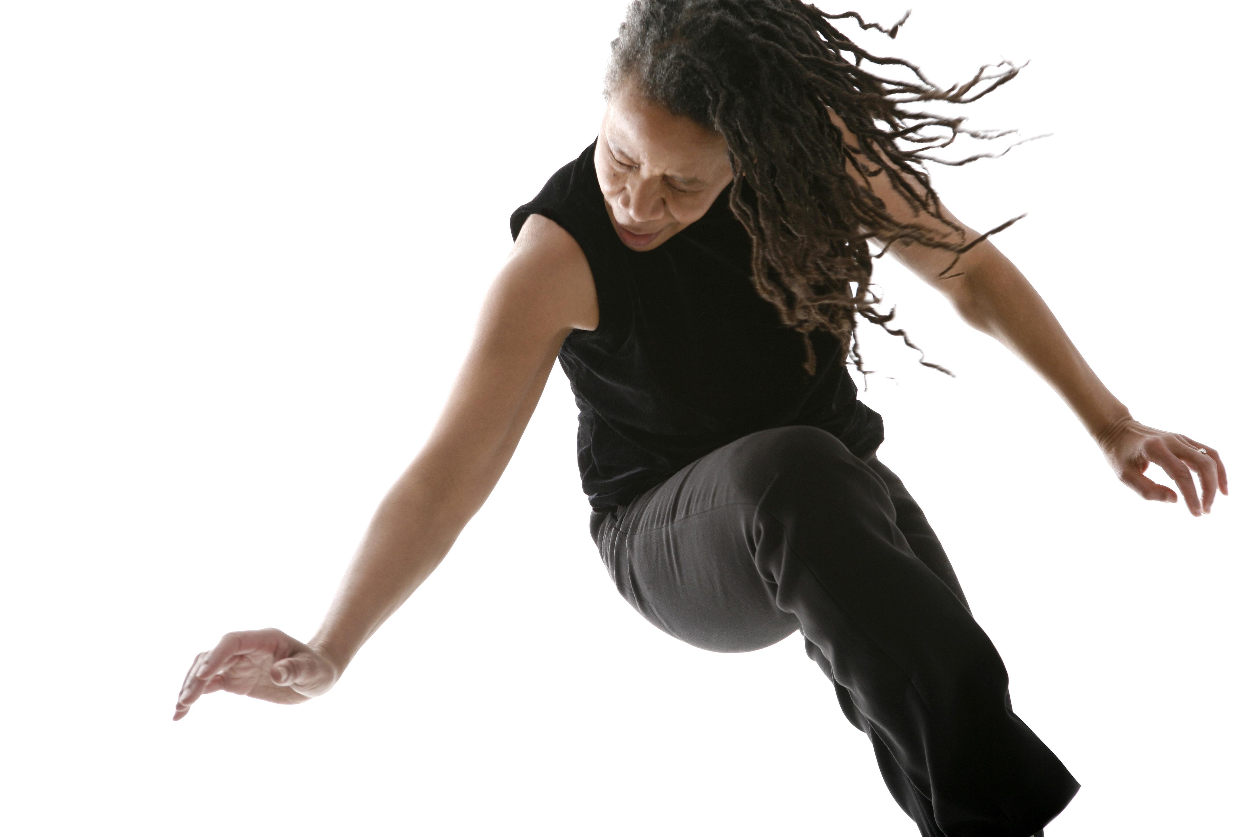 A woman pivots on her left leg, raising her right knee and ducking her head, her long dreadlocks swinging forward her as she turns.