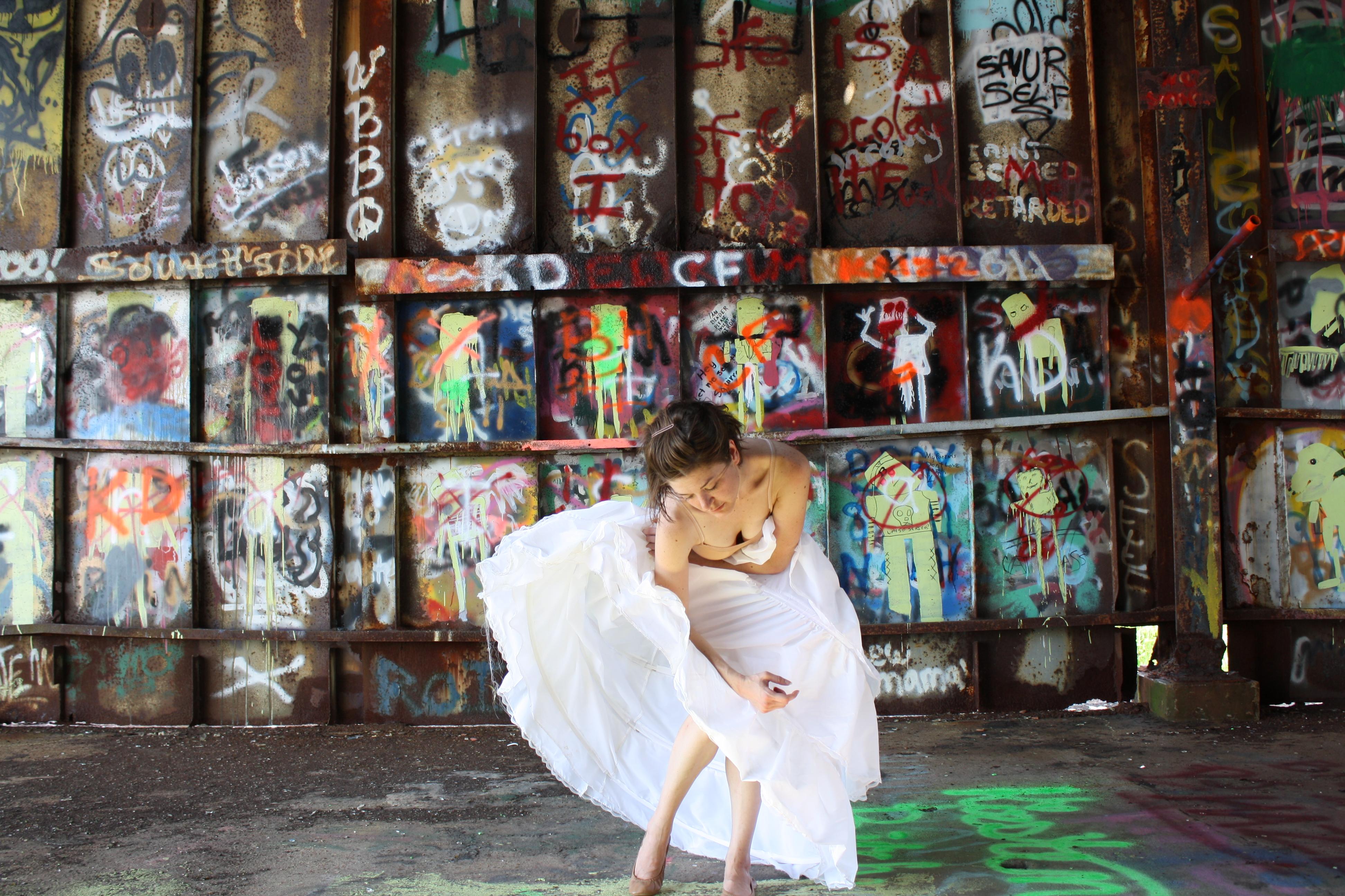 In an urban space filled with graffiti art a light-skinned brunette in a flowy white skirt gracefully bends her body forward in a dance-like motion