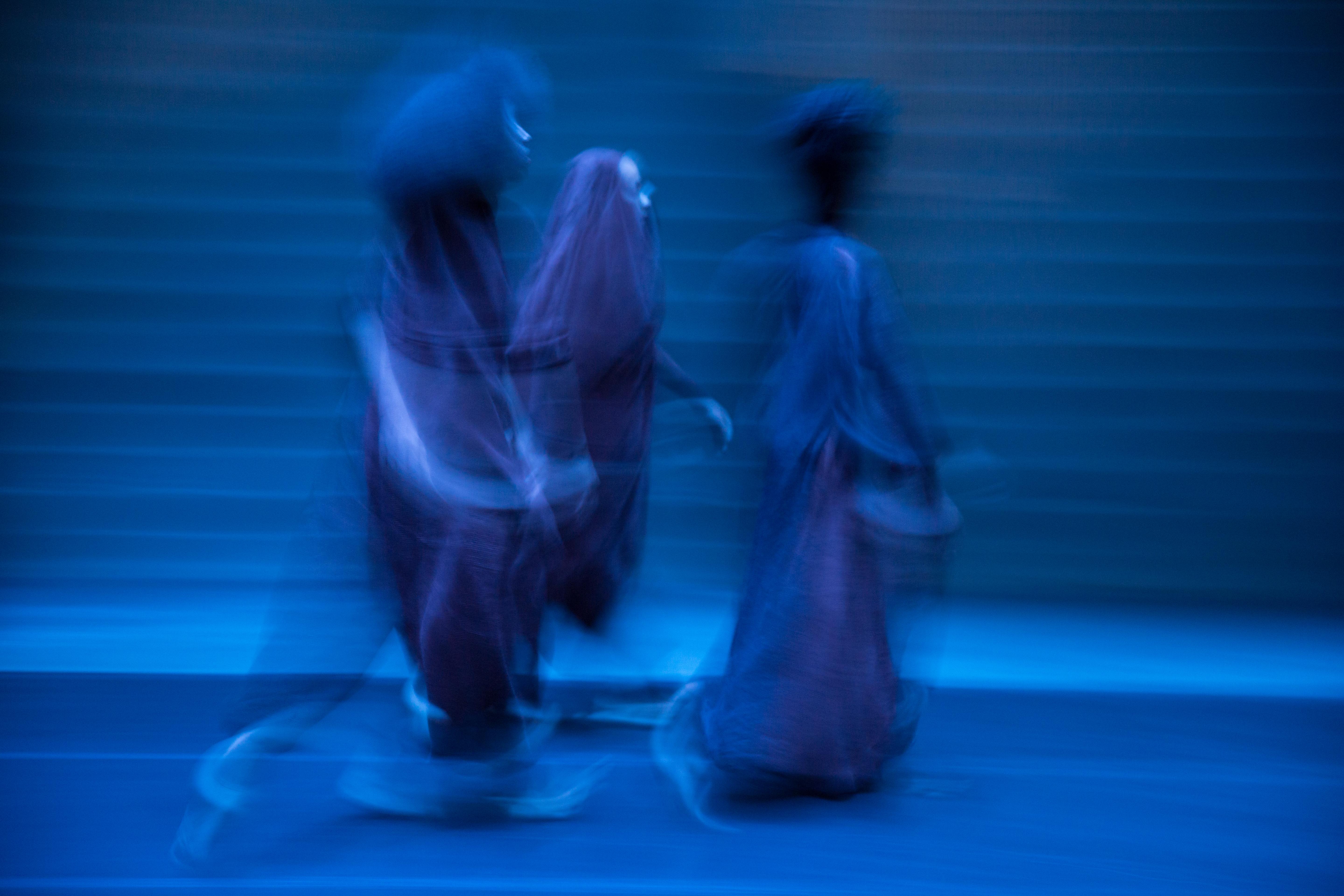 In this blue-toned photograph, three blurry, ghostlike figures in long dresses or robes walk toward the right side of the frame.