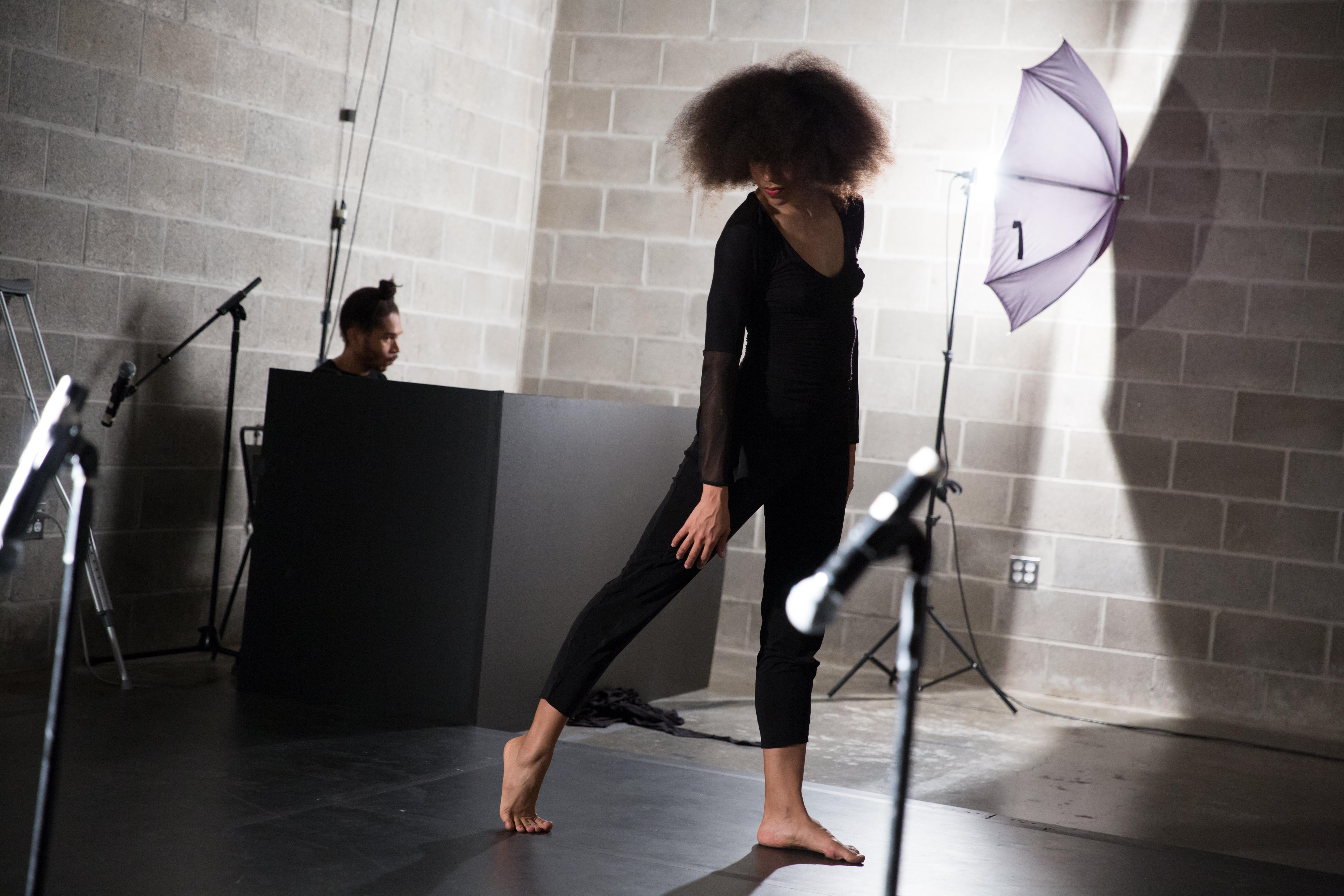 In a cement block–walled space, set like a photography studio with lights and sound equipment, a medium dark–skinned dancer in black clothes poses. Looking down, their face is obscured by curly brown hair.