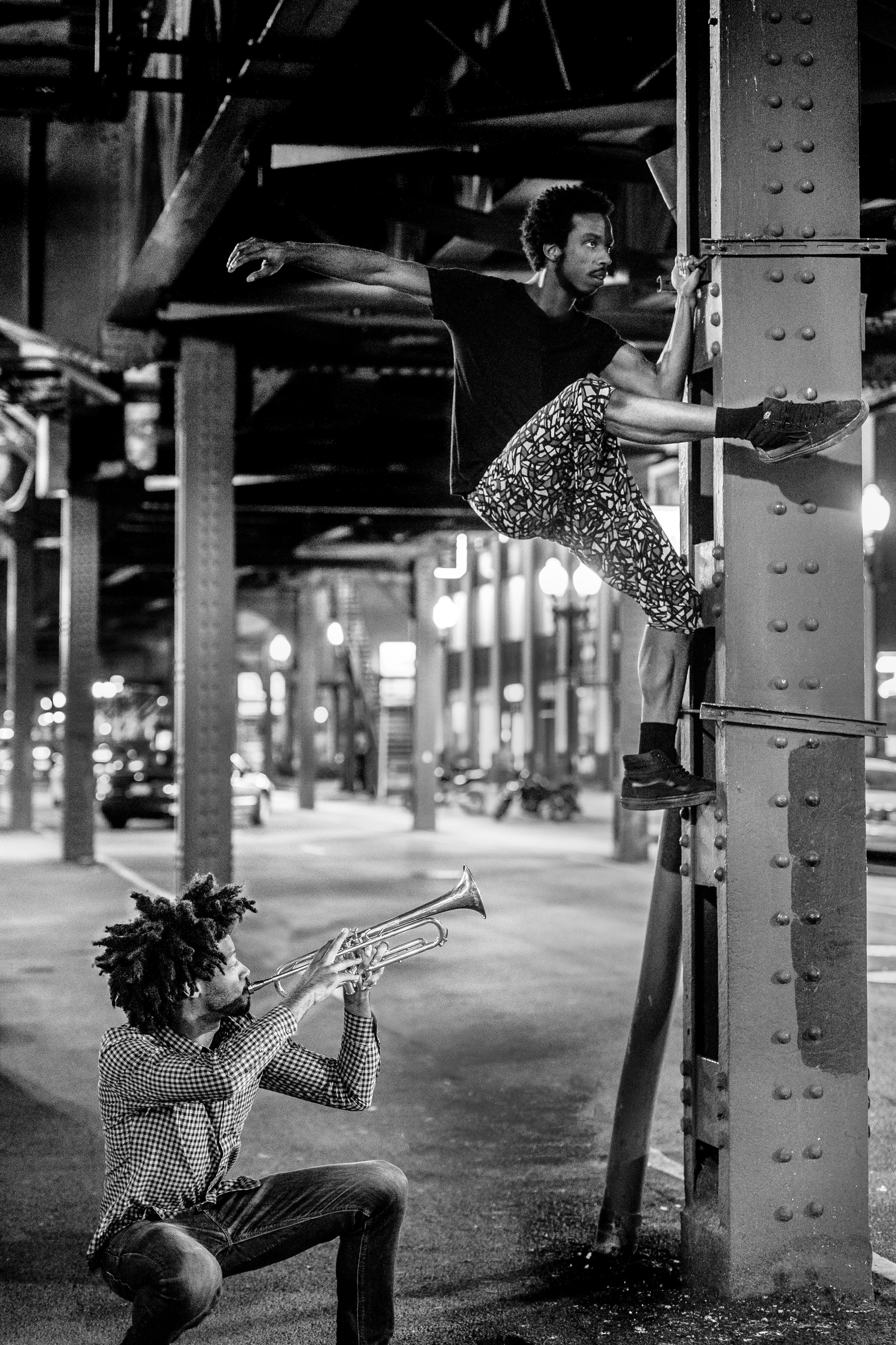 under elevated train tracks a crouching man plays a trumpet while another poses mid-climb up a support column