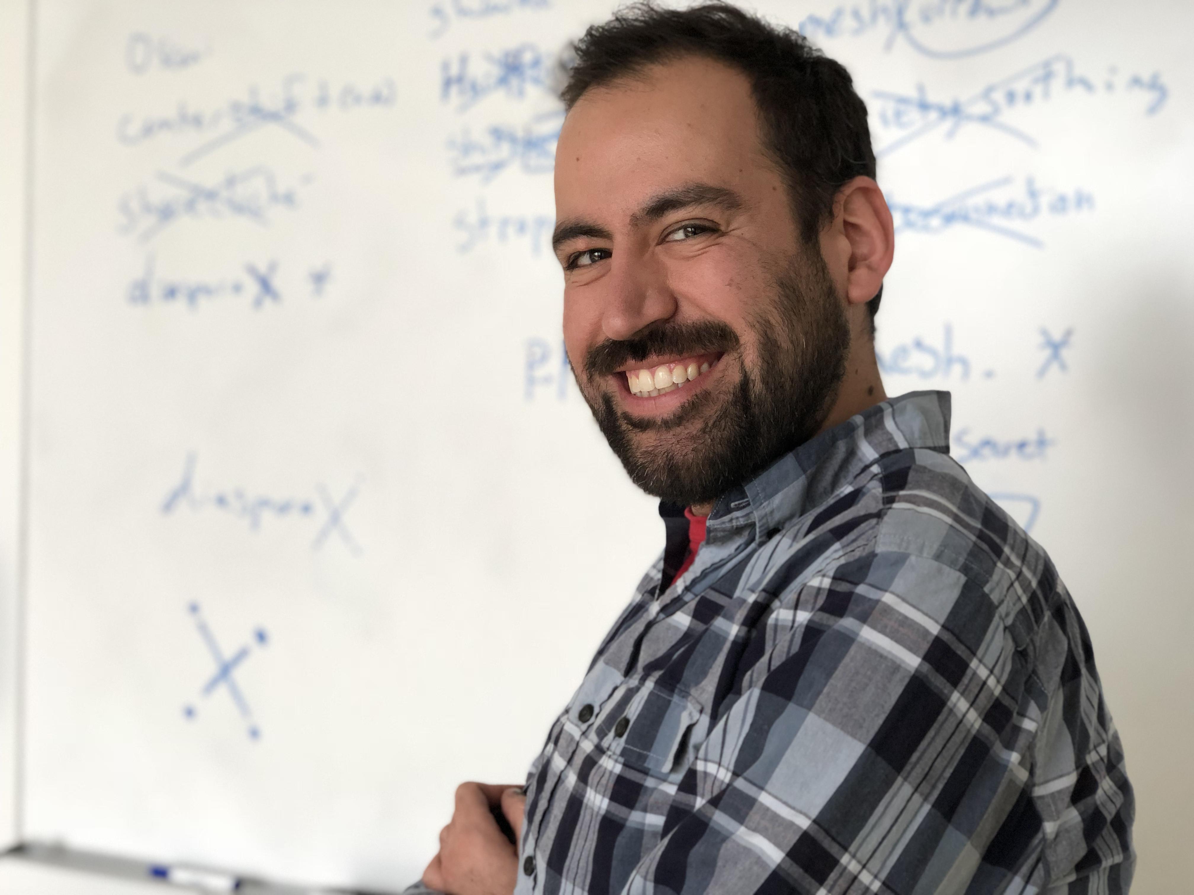 A man in a plaid shirt crosses his arms and faces the viewer. He stands in front of a whiteboard with lists of words, some crossed out.