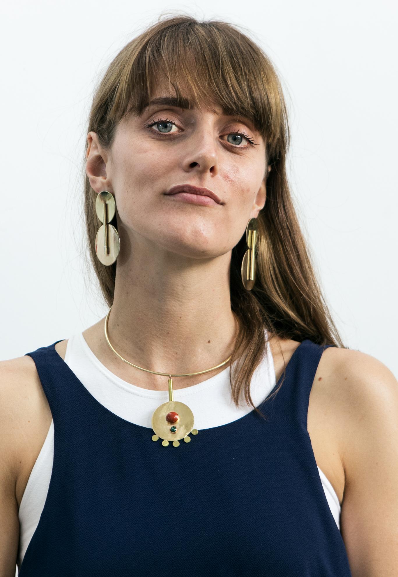 A feminine person with long brown hair and bangs wears large, geometric statement earrings and a bold necklace.