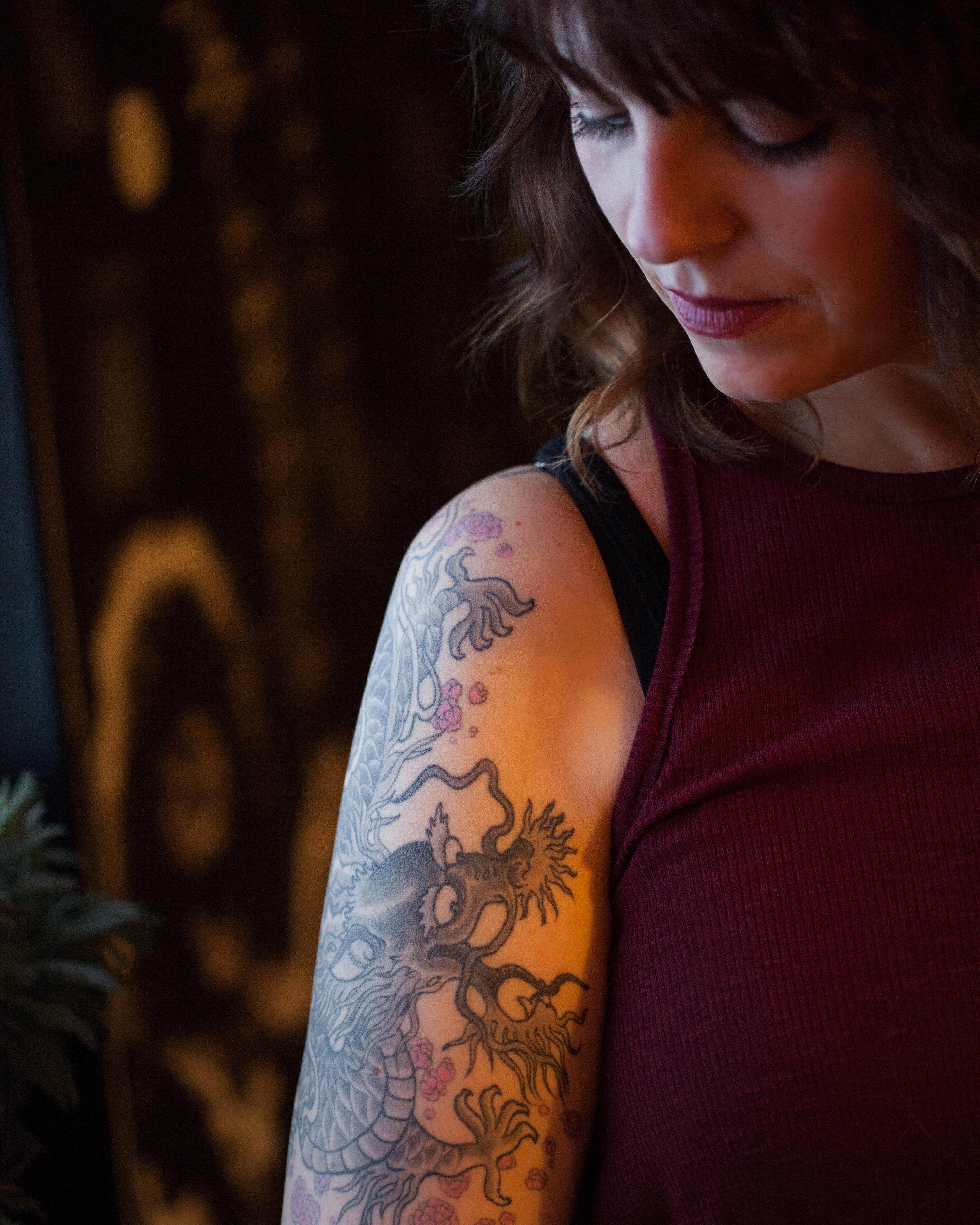 In an artistic portrait, a feminine person with curled brown hair looks down and to their right. Their exposed arm has a large, prominent tattoo of a Chinese-style dragon.