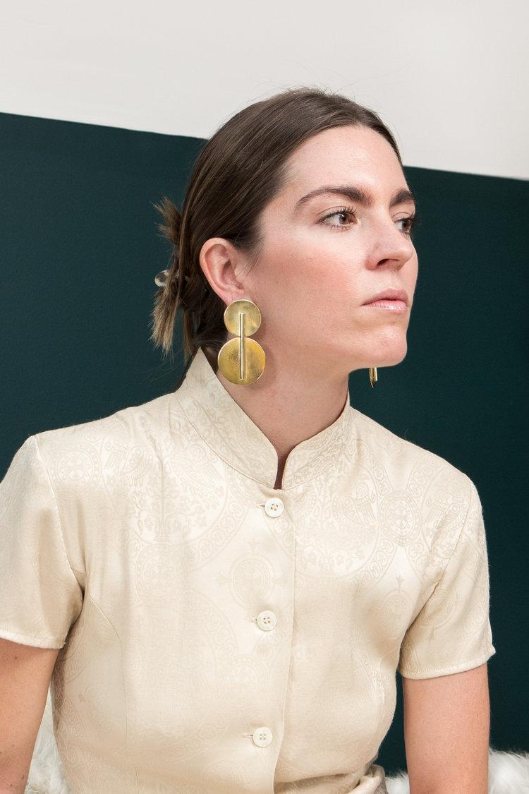 A light-skinned person models a pair of large gold, geometric statement earrings.