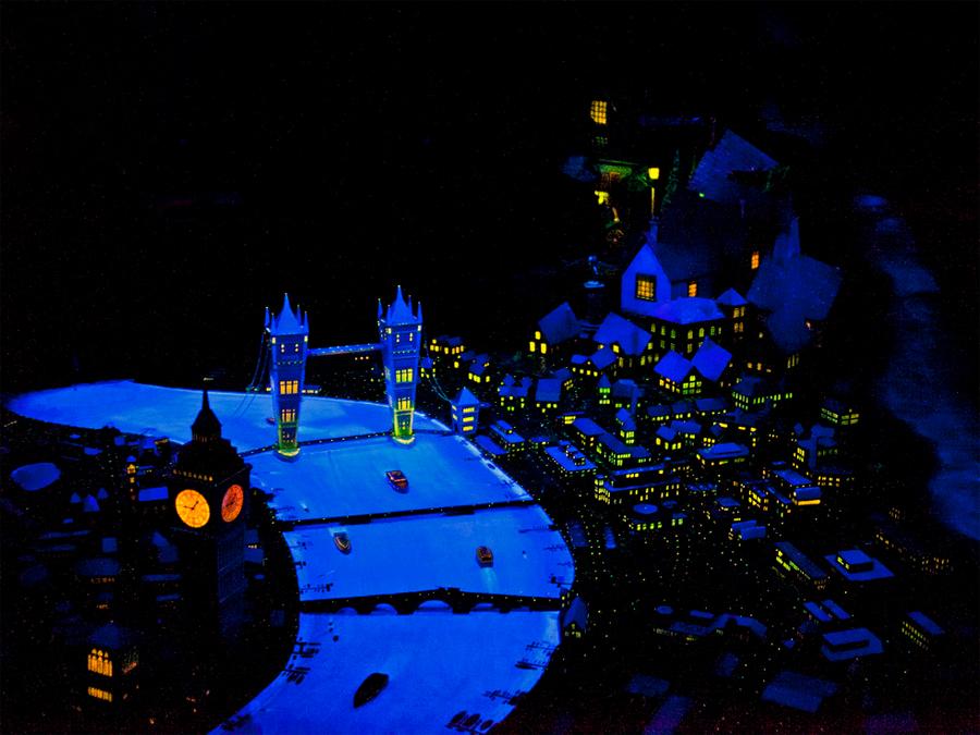 A model reproduction of a cityscape at night is intersected by a river which glows with blue light alongside the warm glowing windows of urban buildings.