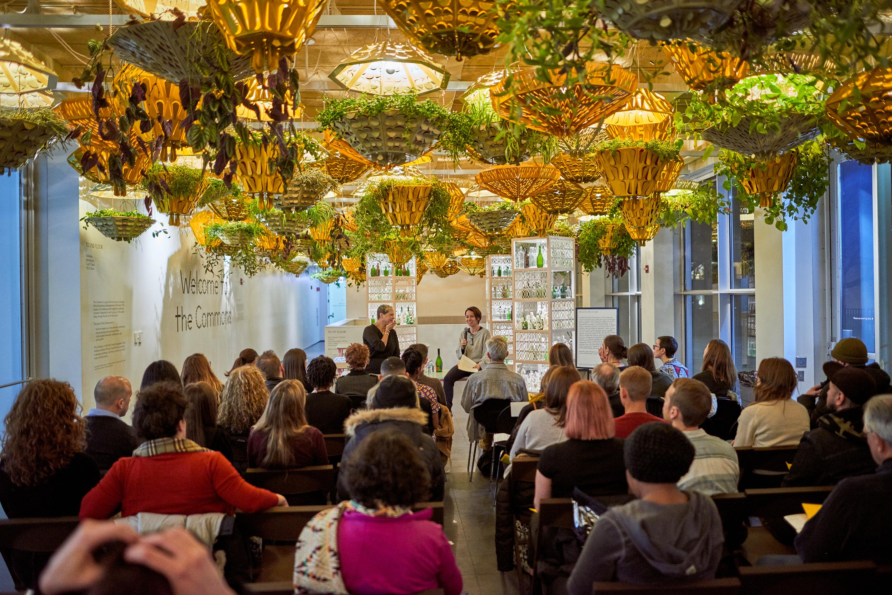seated listeners gather; the space's ceiling is a dense tapestry of green hanging plants in unique golden vessels
