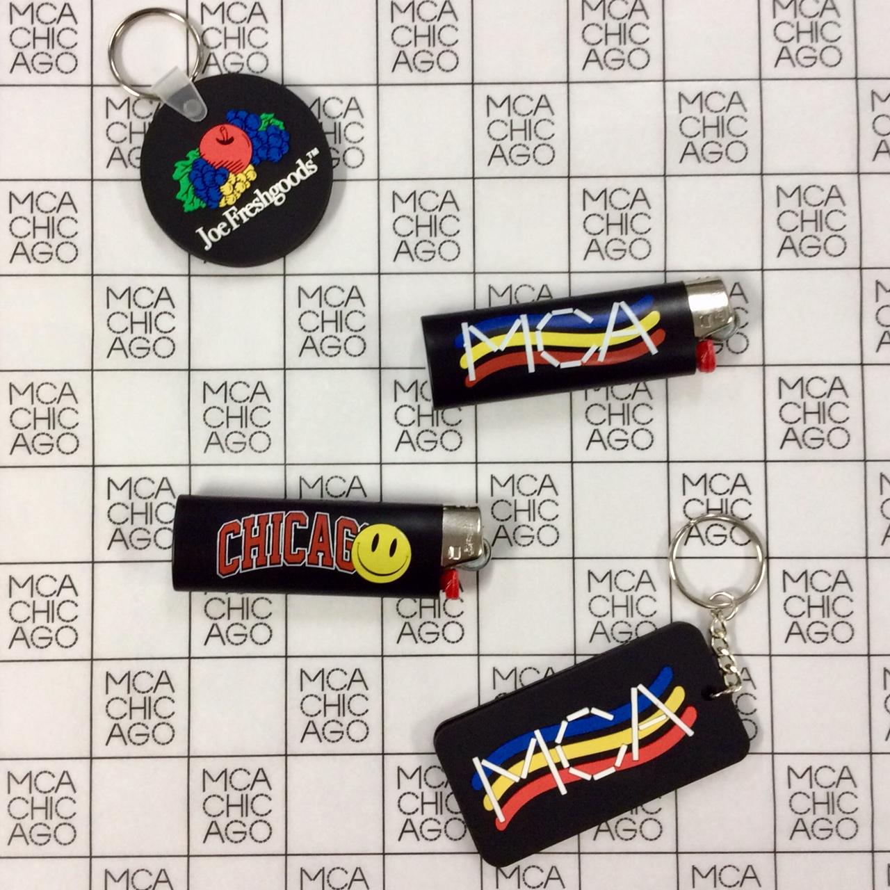 A Joe Freshgoods key chain, MCA-branded lighter and key chain, and a Chicago lighter sit on a black-and-white grid of MCA Chicago logos.