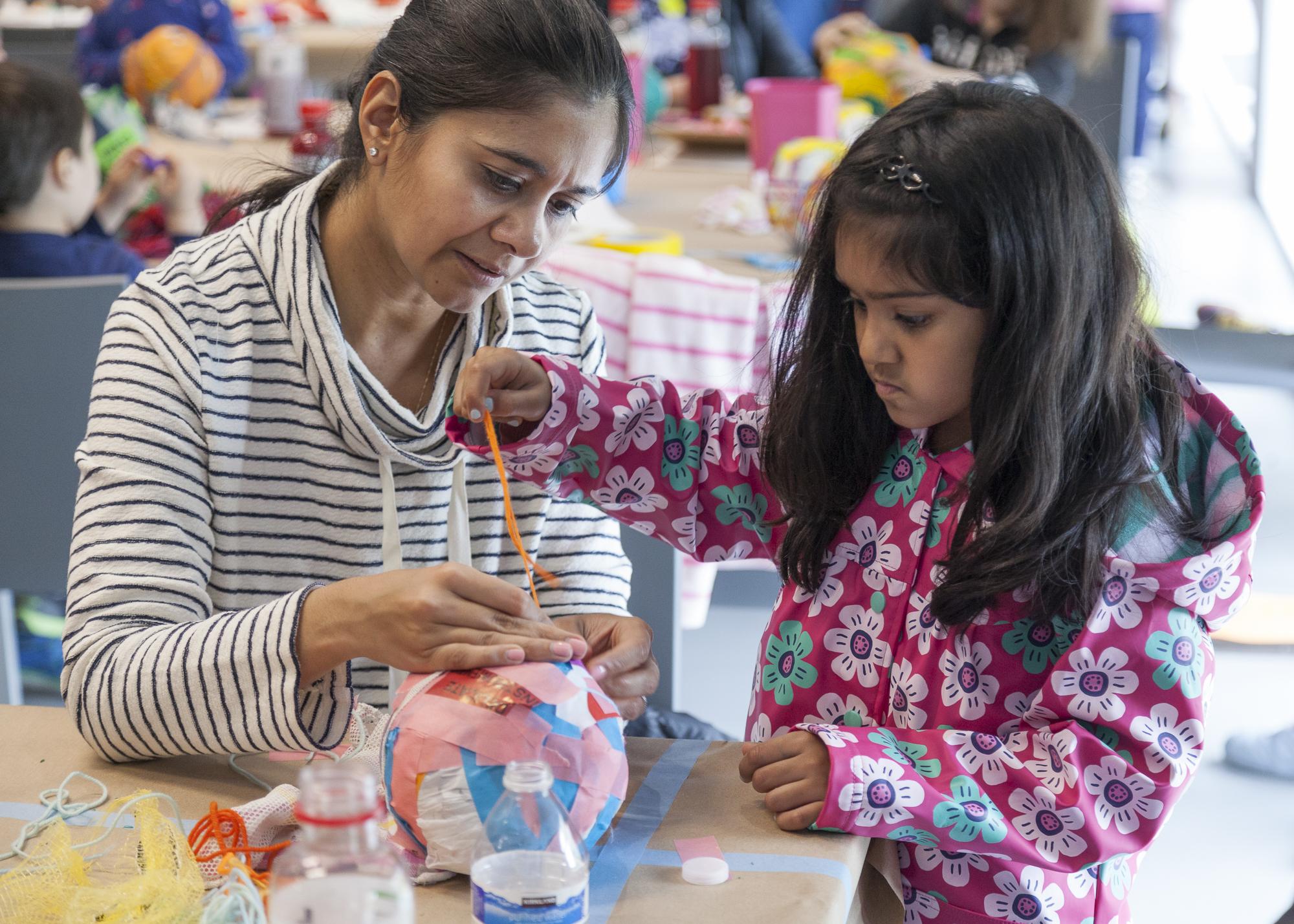 A child in a pink floral coat wraps an orange string around a colorful paper ball. An adult holds the string in place, assisting the child.