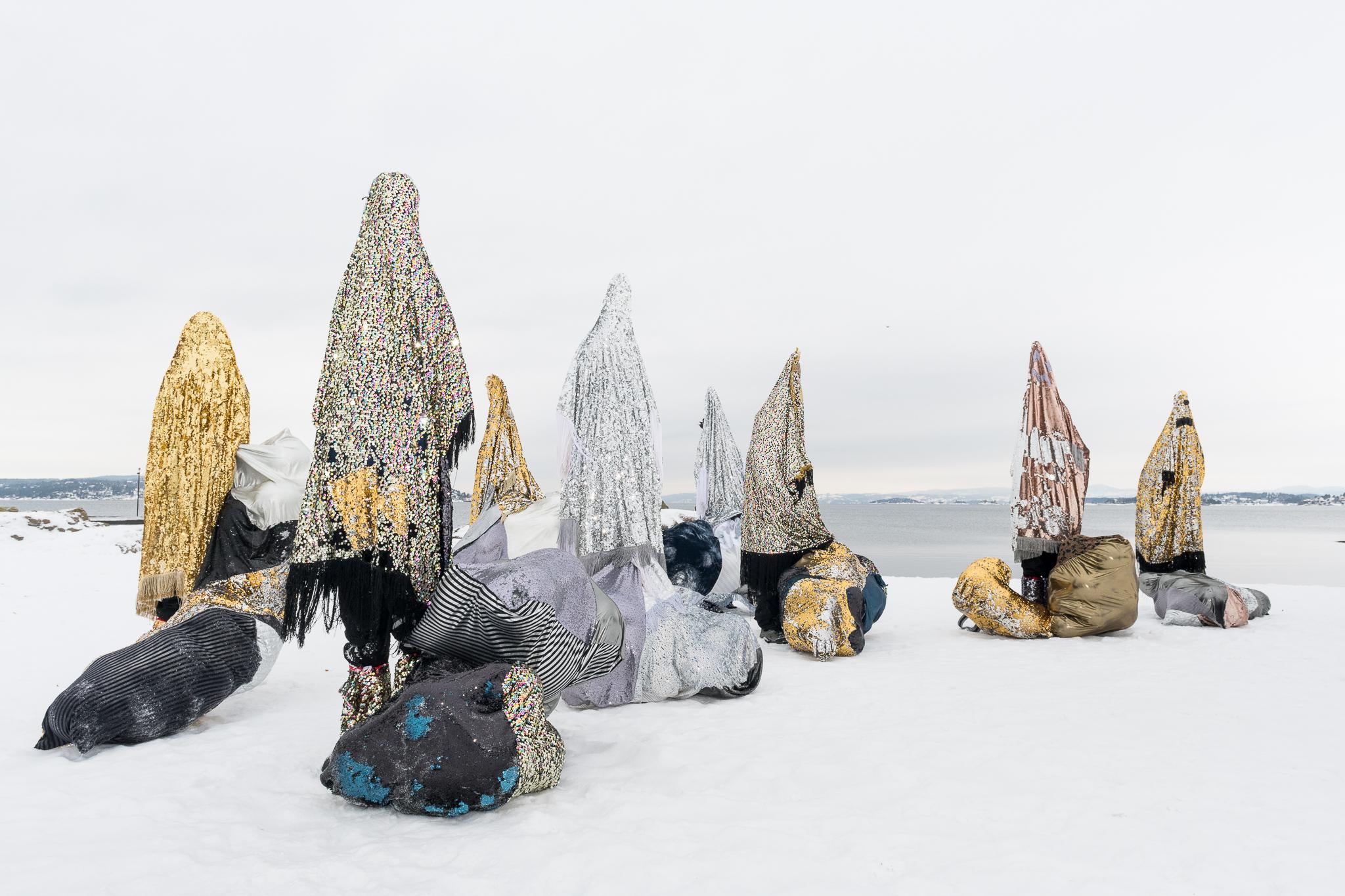 Figures draped in sequin fabric of metallic colors stand against a snowy scene. Some of the figures have pointed tips, and bulbous shapes in similar fabric are on the ground.