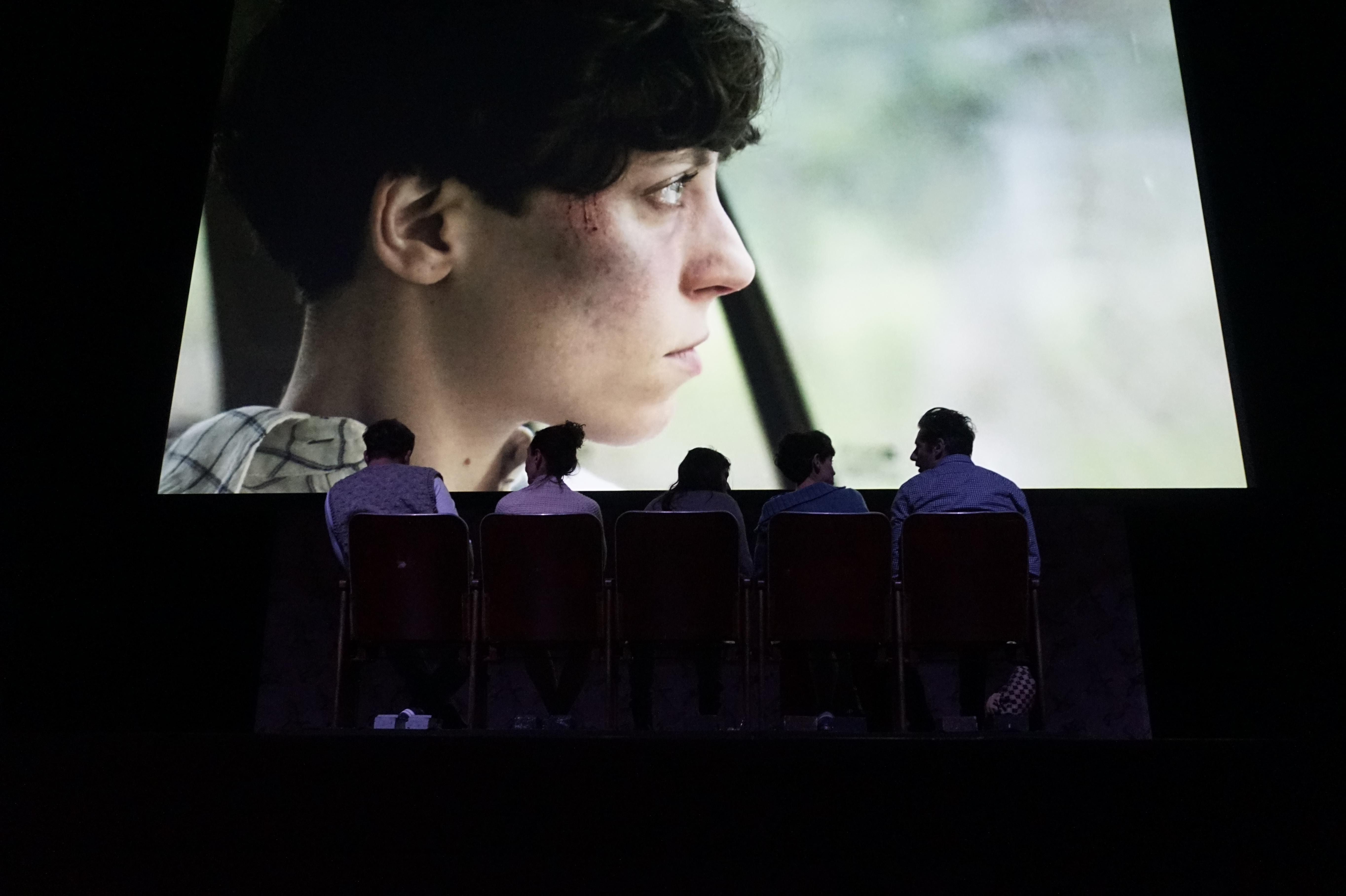 Five people in a darkened space sit on chairs with their backs to the audience; in front of them is a giant projected profile of a short-haired person looking to the right.