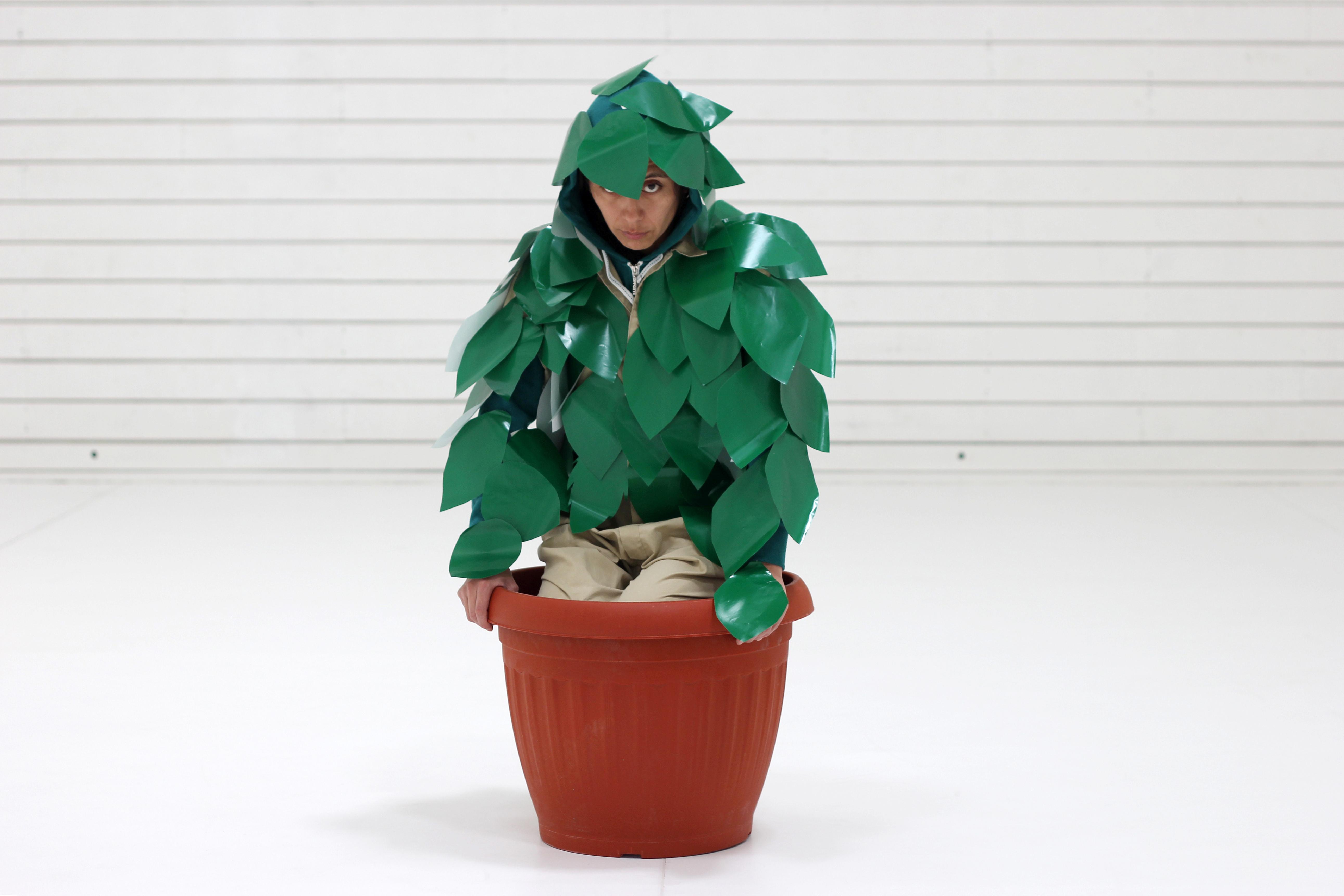 A person covered in shiny cut-out leaves squats in a large planter.