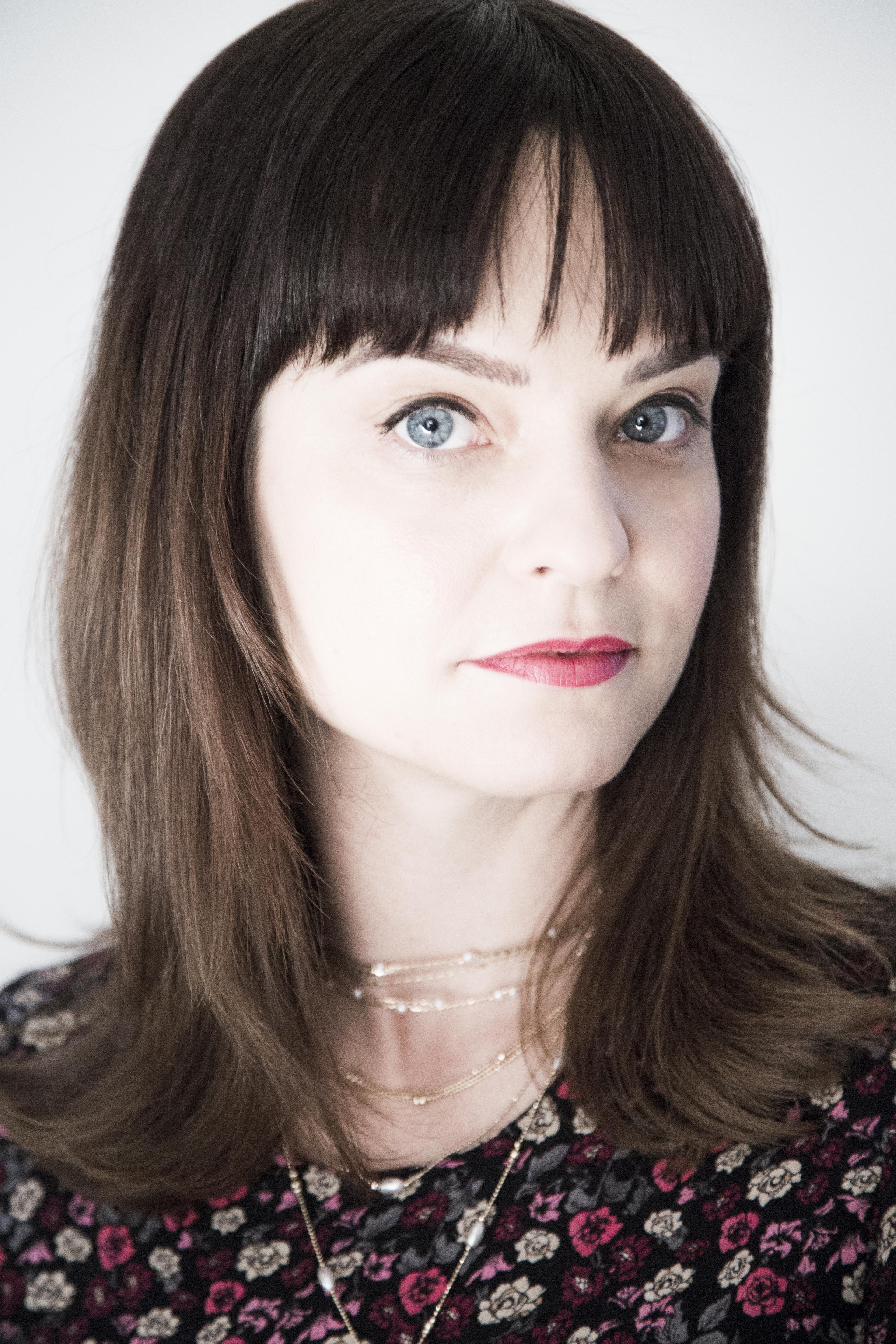 A headshot photo shows a light-skinned woman with long dark hair and blue eyes. She wears gold necklaces and a flower-patterned dress.